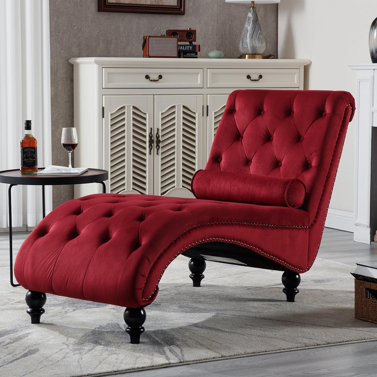 Close-up of a red tufted S-shaped chaise angled left with bolster pillow, silver nailhead trim and turned black legs.