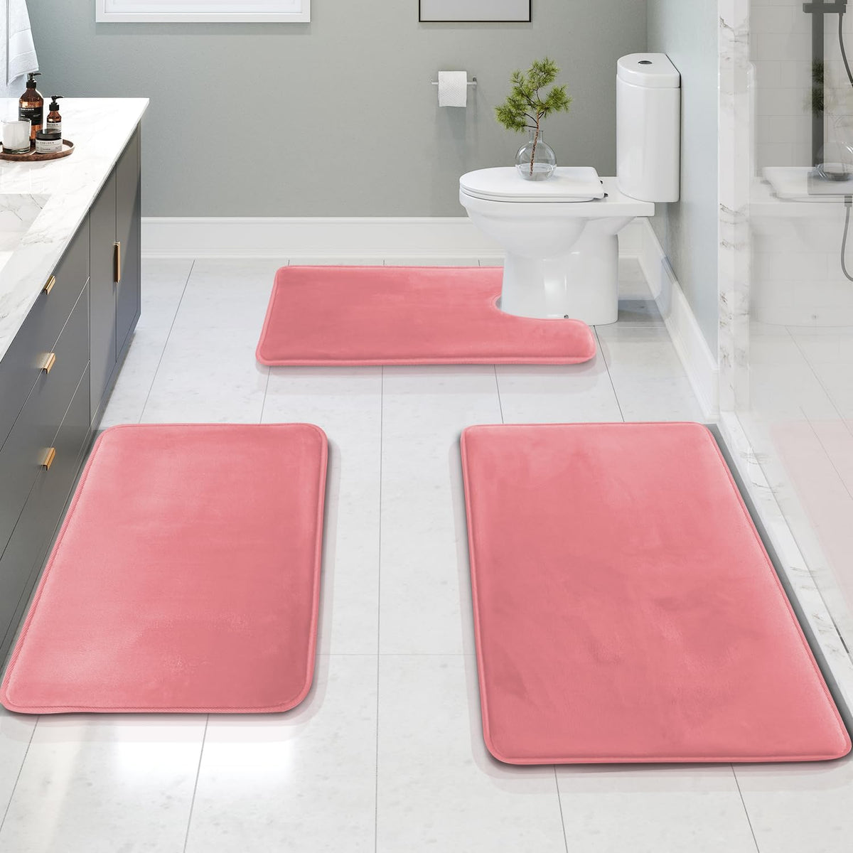 Bathroom scene with three pink memory-foam bath mats arranged on a tiled floor around a toilet and vanity.