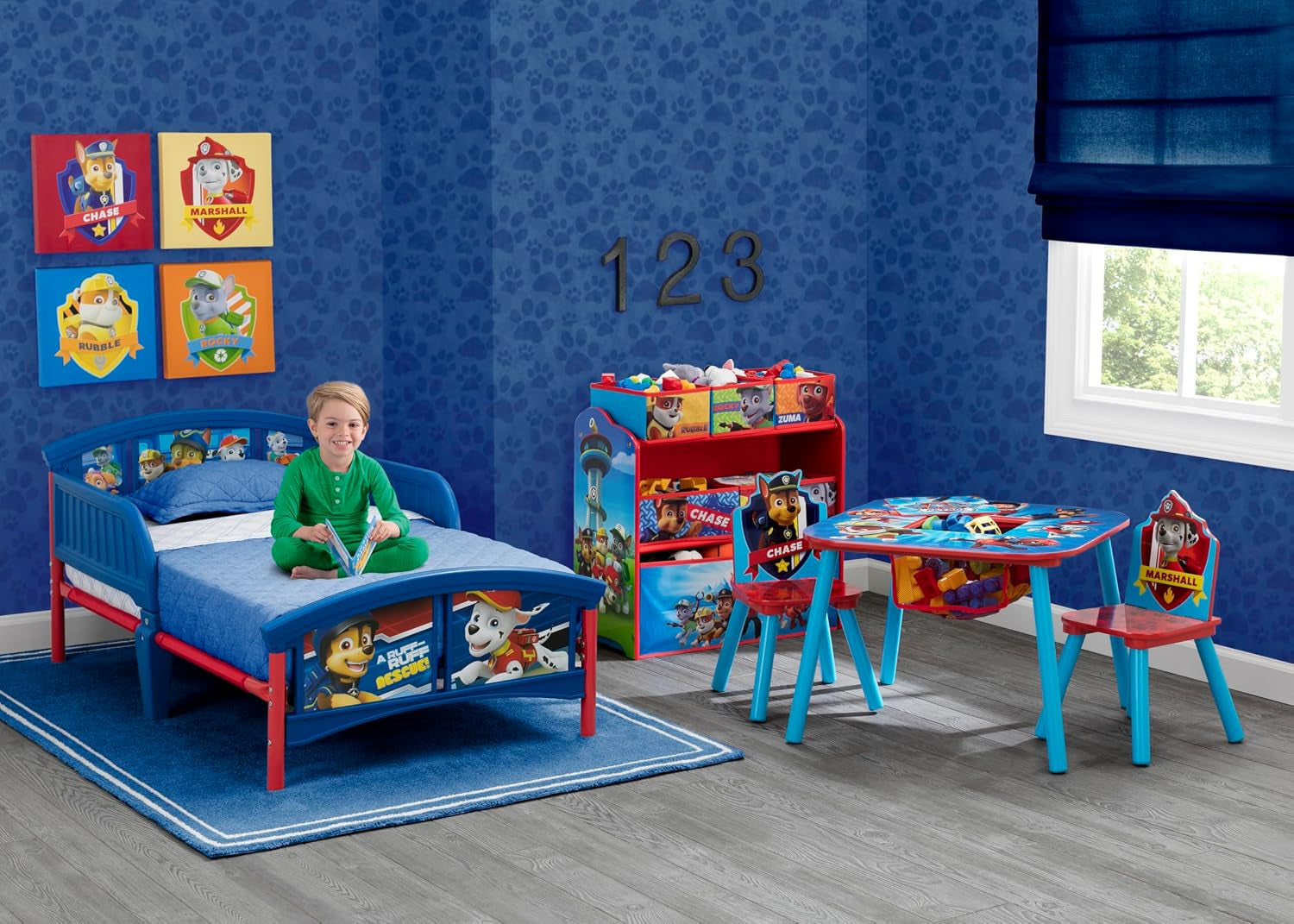Child sitting on the Paw Patrol-themed toddler bed in a blue playroom with matching table, chairs, and storage.