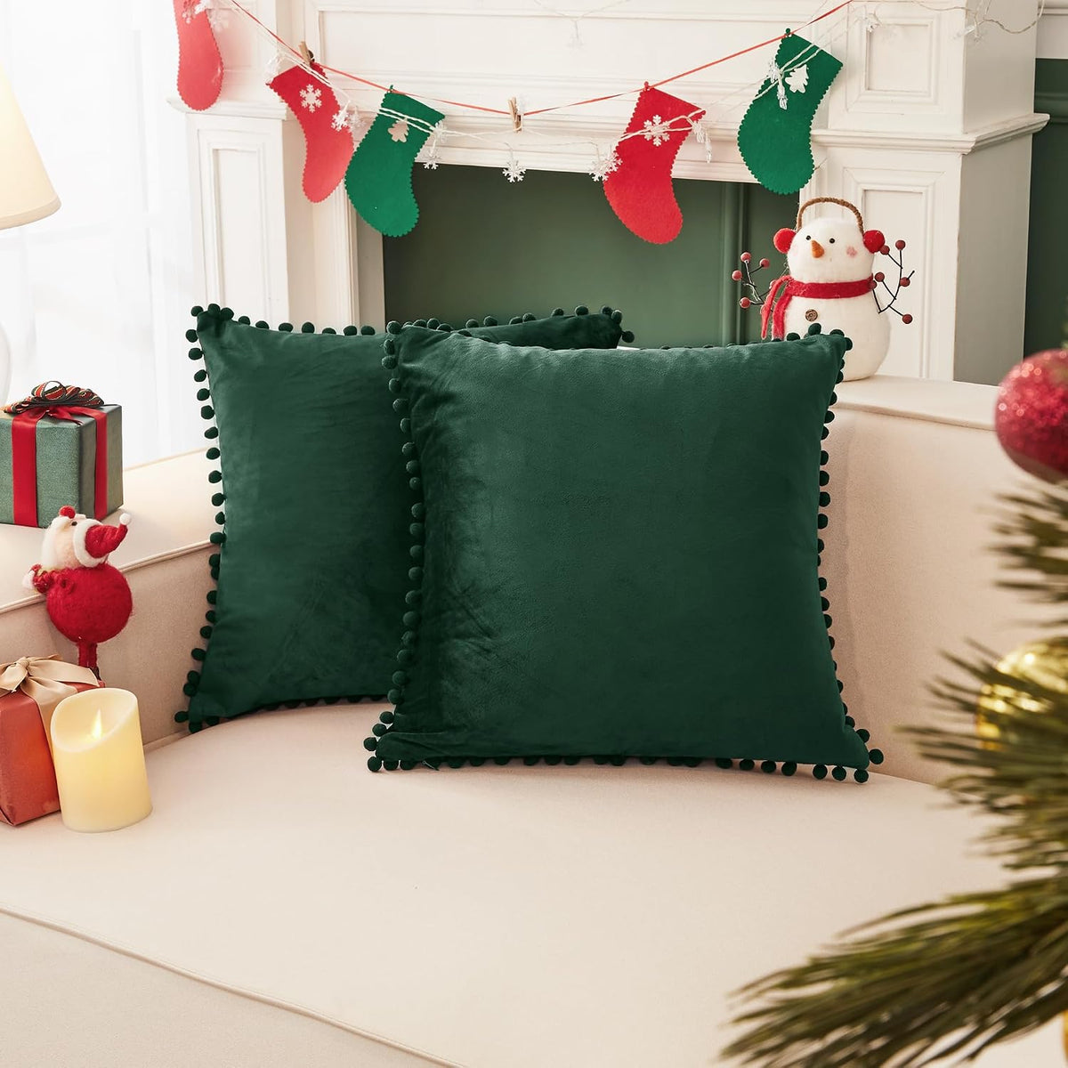 Two dark green square pillow covers with pom-pom trim on a couch beside holiday gifts and candles