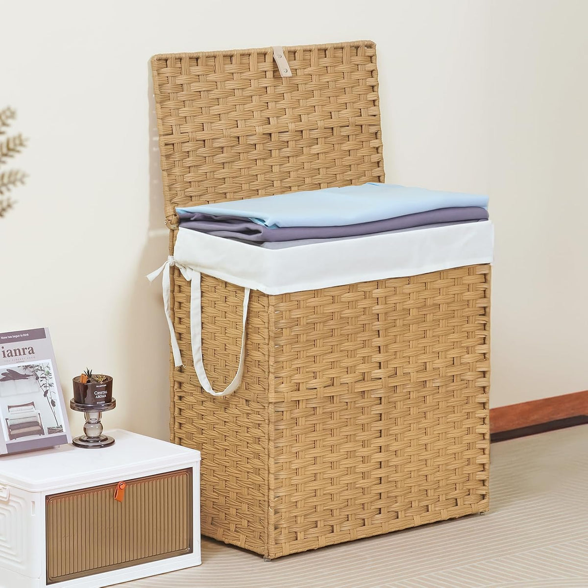 Tall tan rattan hamper with lid propped open, white liner showing and two folded sheets stacked on top.