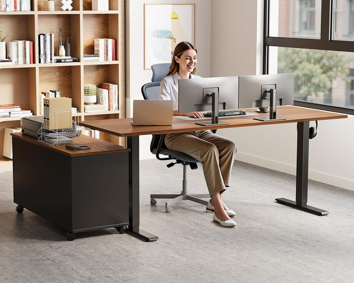 Woman seated at walnut L-shaped standing desk using dual monitors with the mobile black file cabinet beside the desk