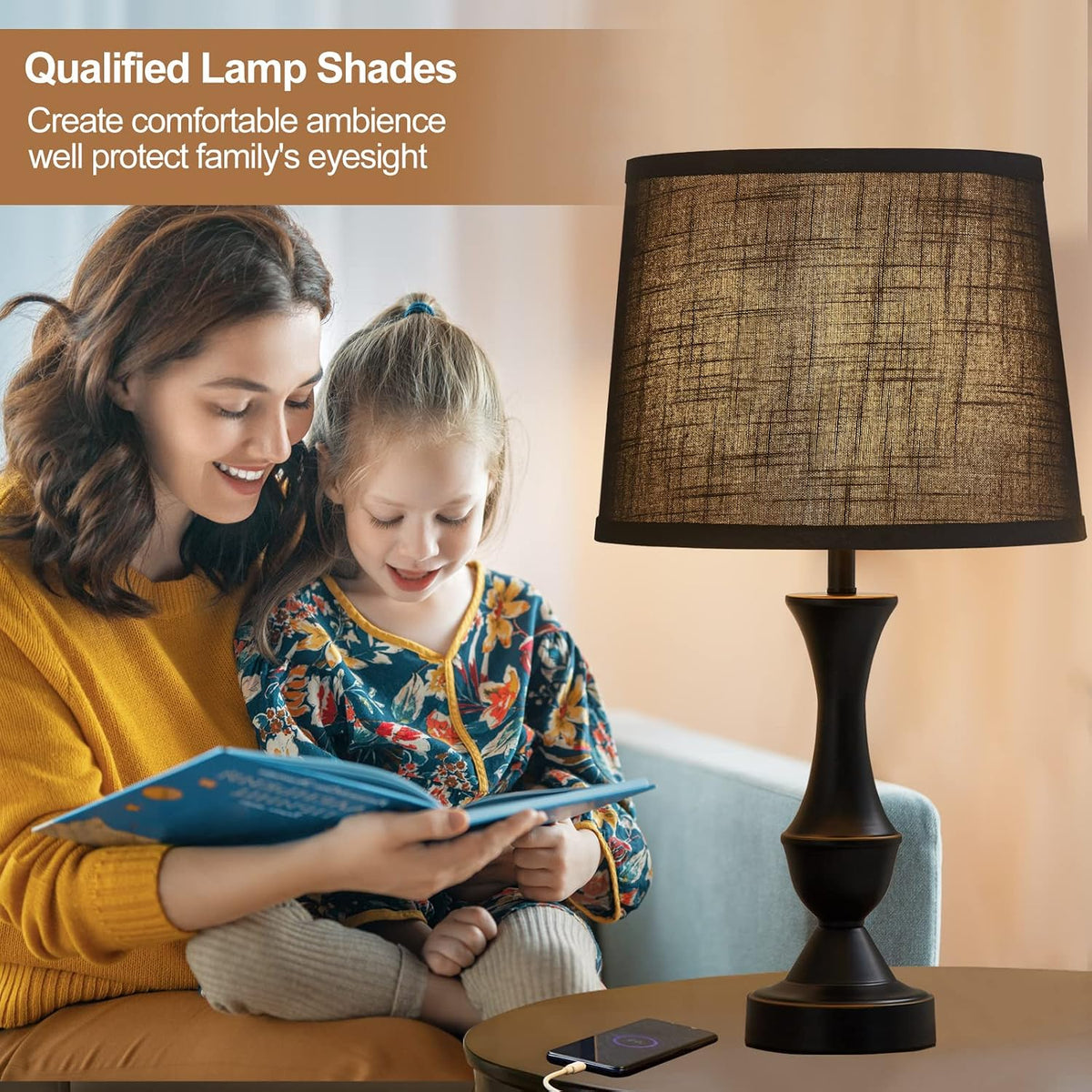 Woman and child reading beside a lit table lamp topped by a dark textured linen shade.