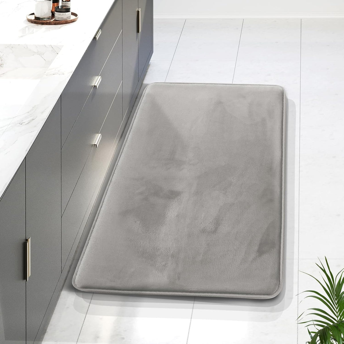 Top-down view of a gray rectangular memory-foam bath mat beside a gray vanity on white tile.