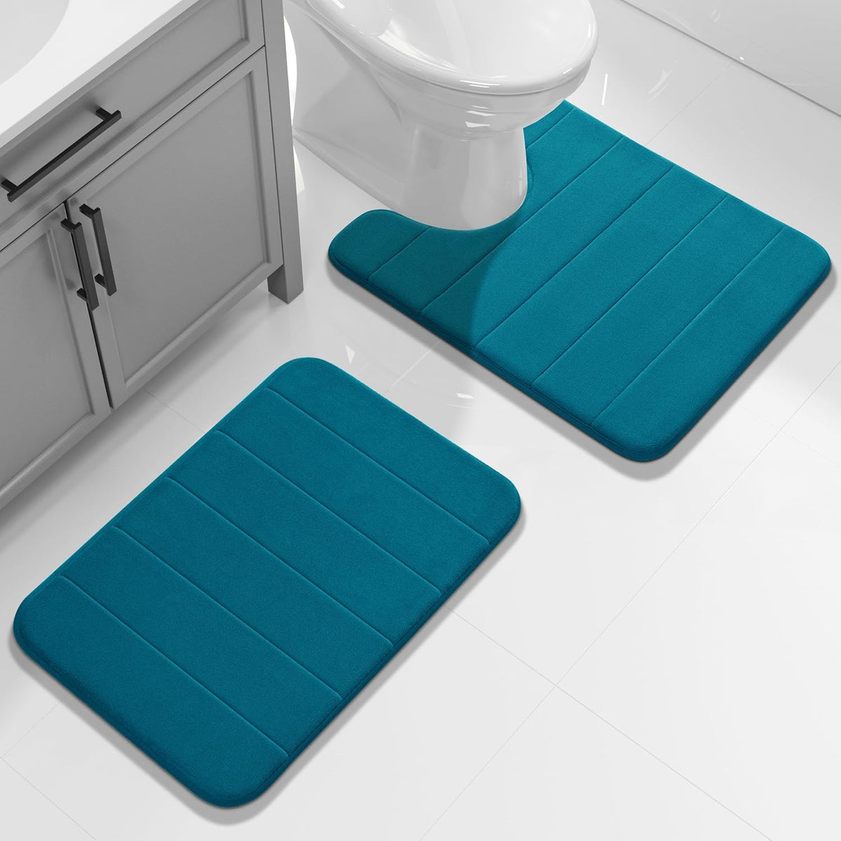 Two deep teal rectangular mats on a white tiled bathroom floor positioned near a cabinet and toilet.