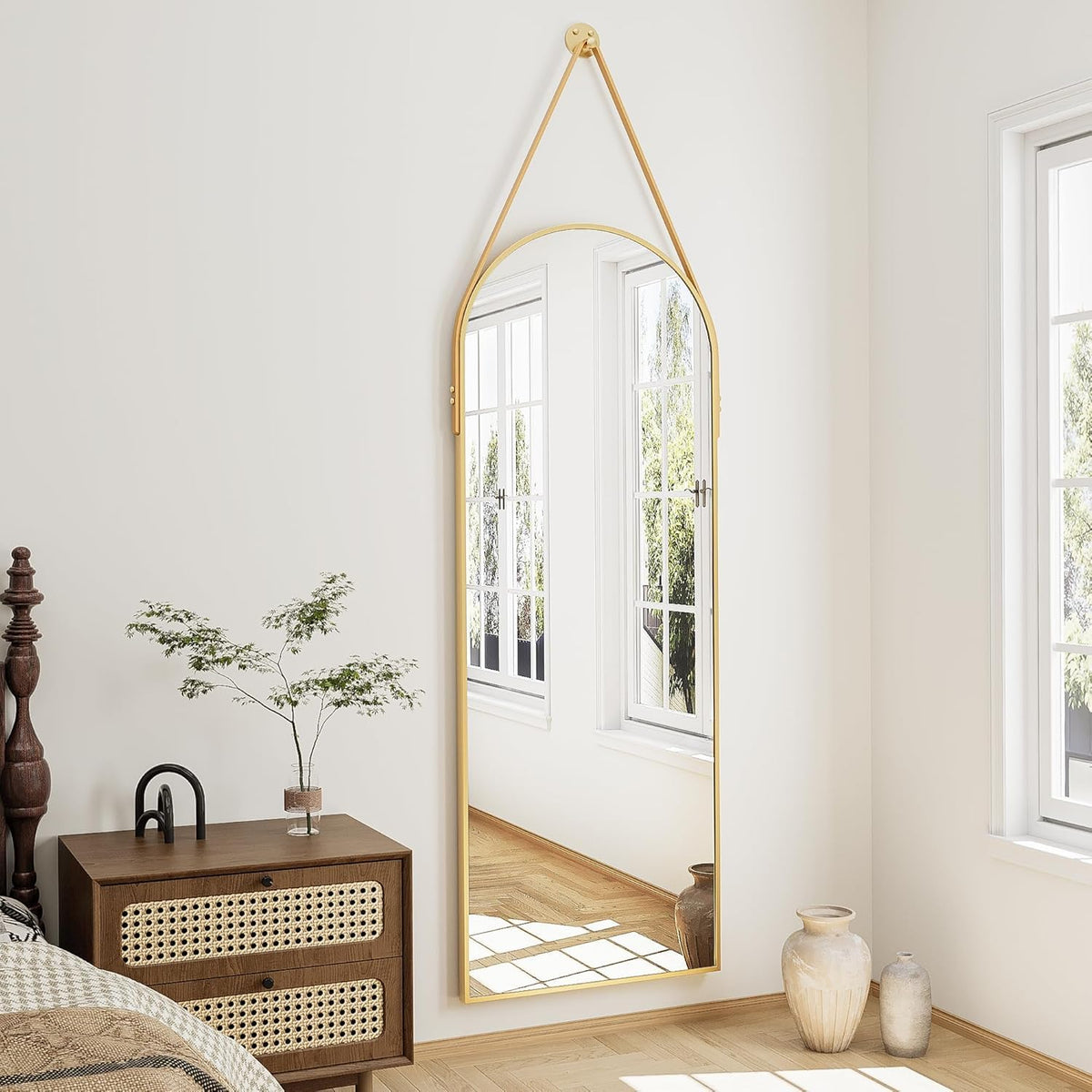 Tall arched mirror with gold frame suspended from a single strap, reflecting bright windows and parquet floor.