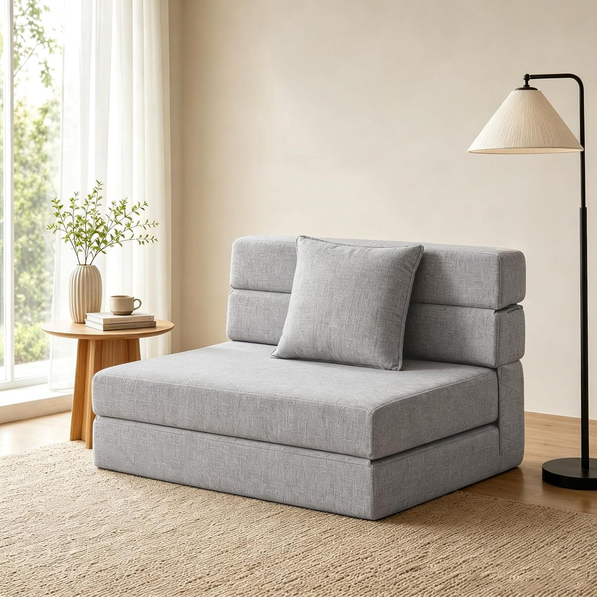 Light gray folding futon sofa bed beside round wooden side table and tall floor lamp