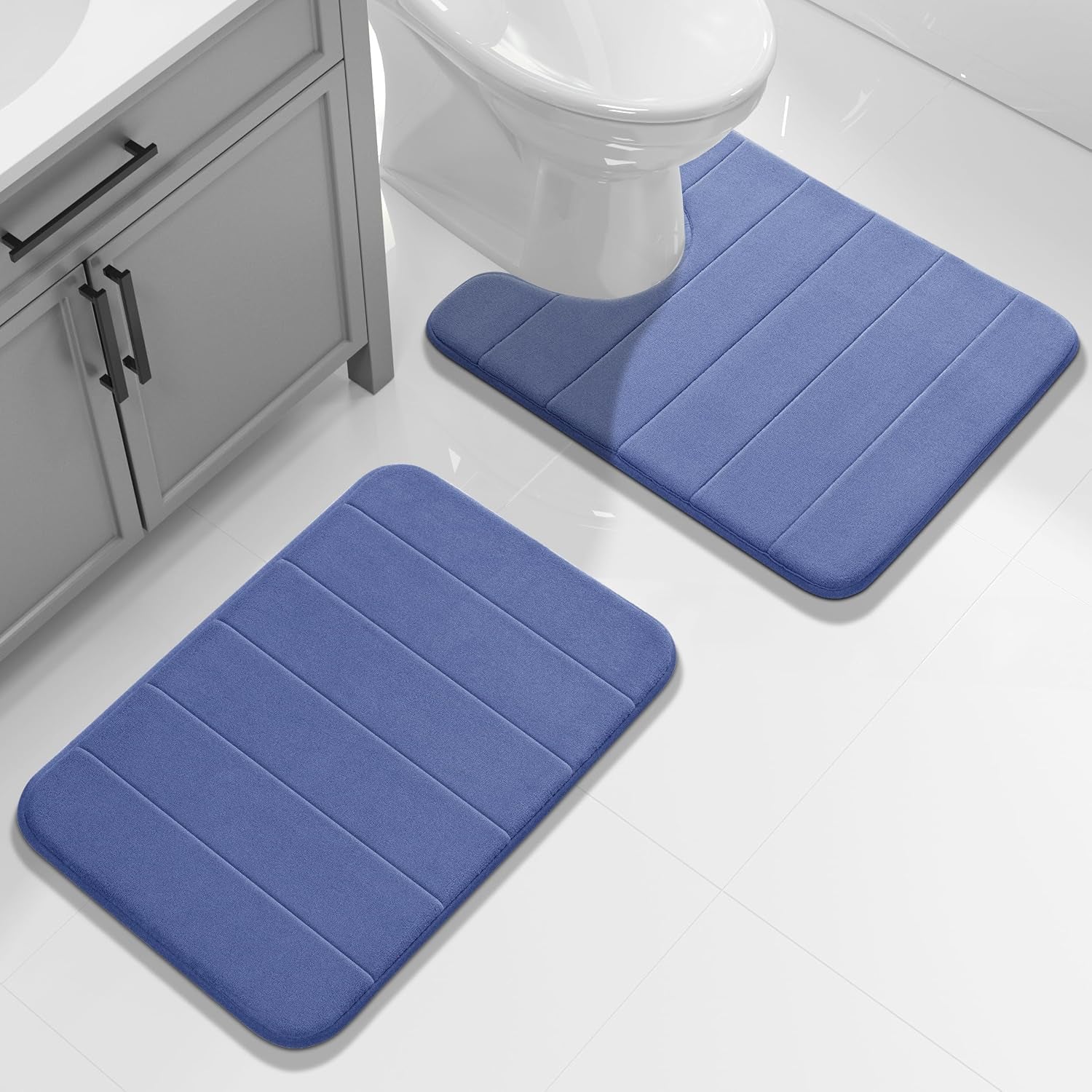 Blue two-piece bath rug set on glossy white tile with rectangular mat and contour toilet mat under the bowl