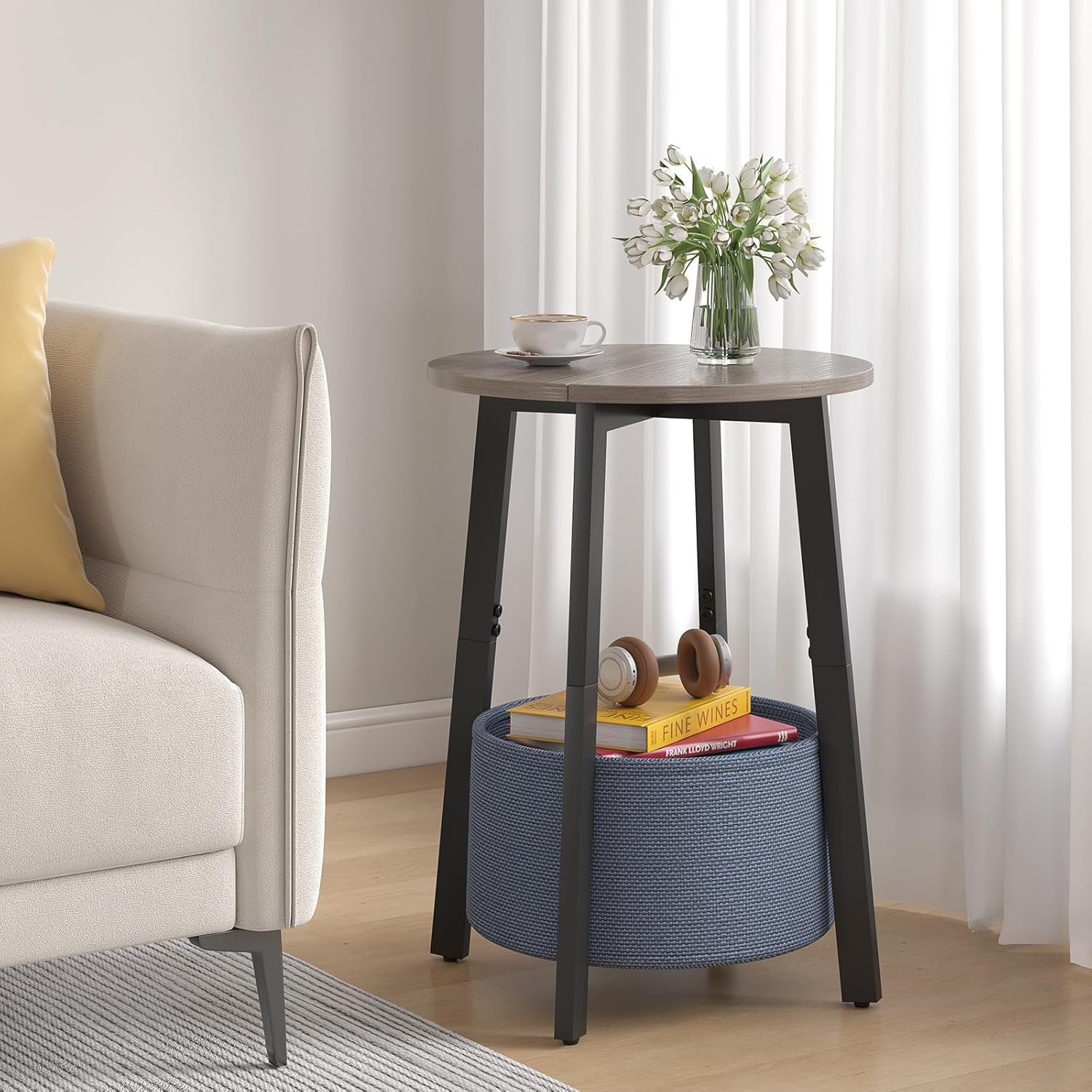Side table next to a sofa with a cup and vase on top and books in the blue woven basket below
