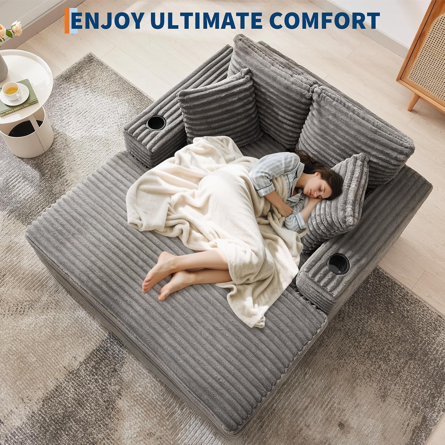 Top-down view of a gray corduroy chaise with cup holders; a woman sleeps under a cream blanket.