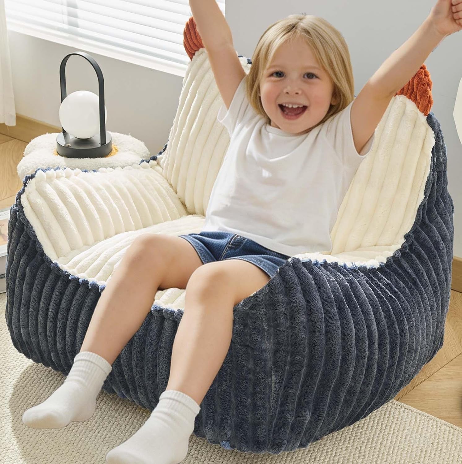 Young child sitting in a ribbed dark blue round bean bag with cream interior next to a small lamp.