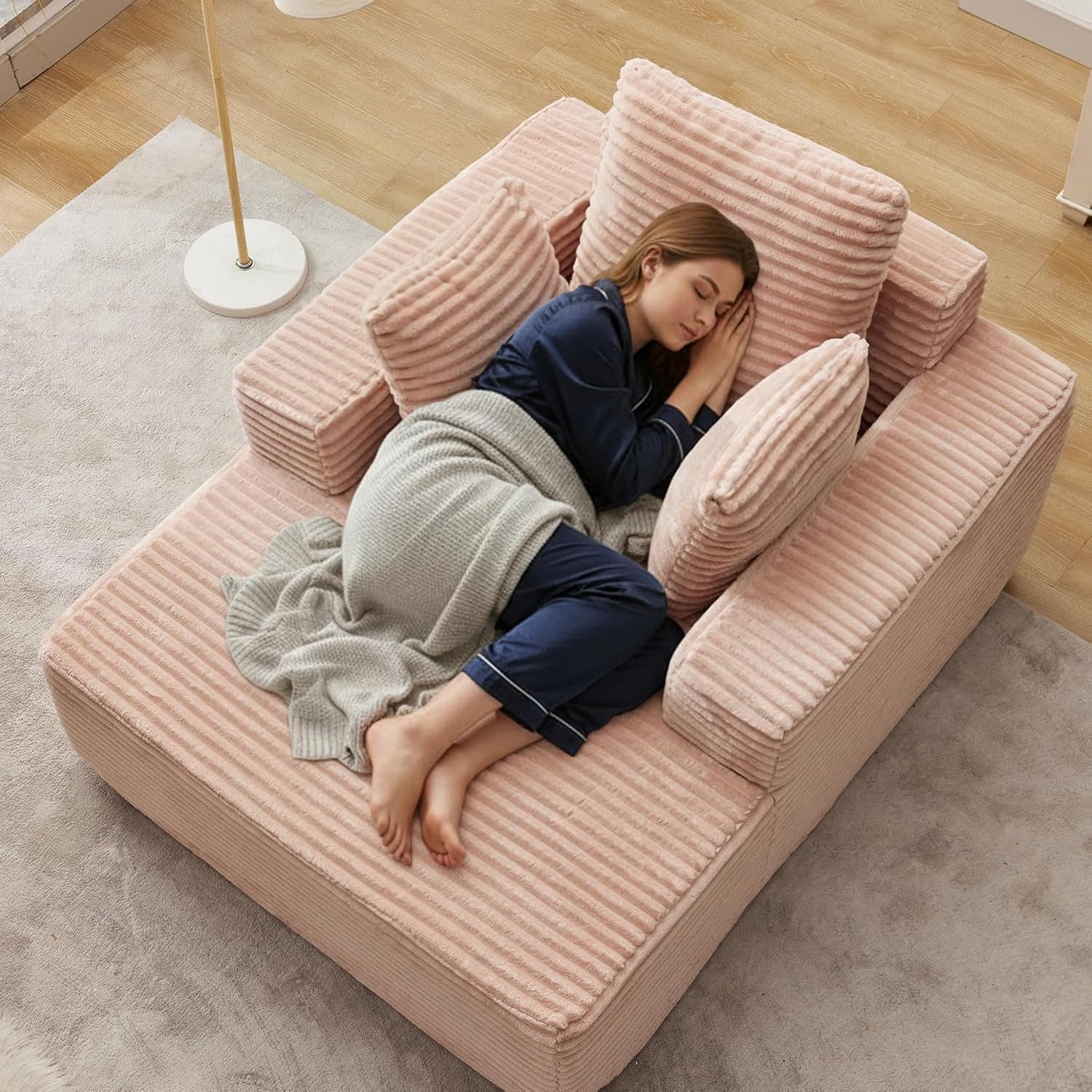 Top-down view of a pink corduroy chaise lounge with three matching pillows; a woman sleeps wrapped in a gray blanket.
