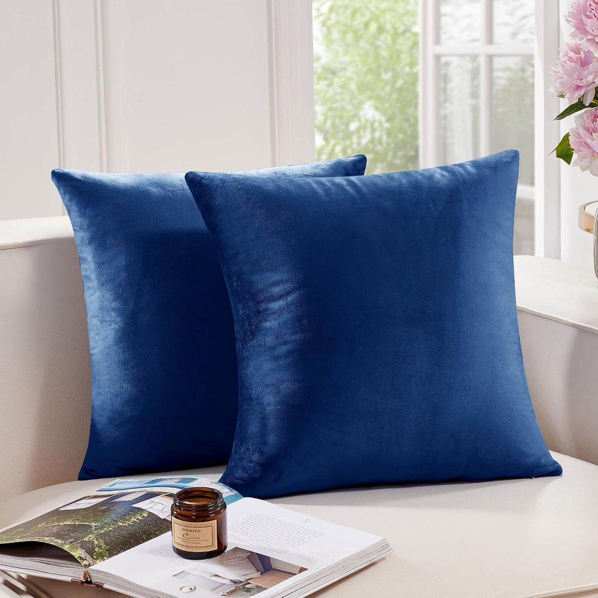 Two deep navy blue square pillow covers leaning on a light sofa beside a candle and open book.