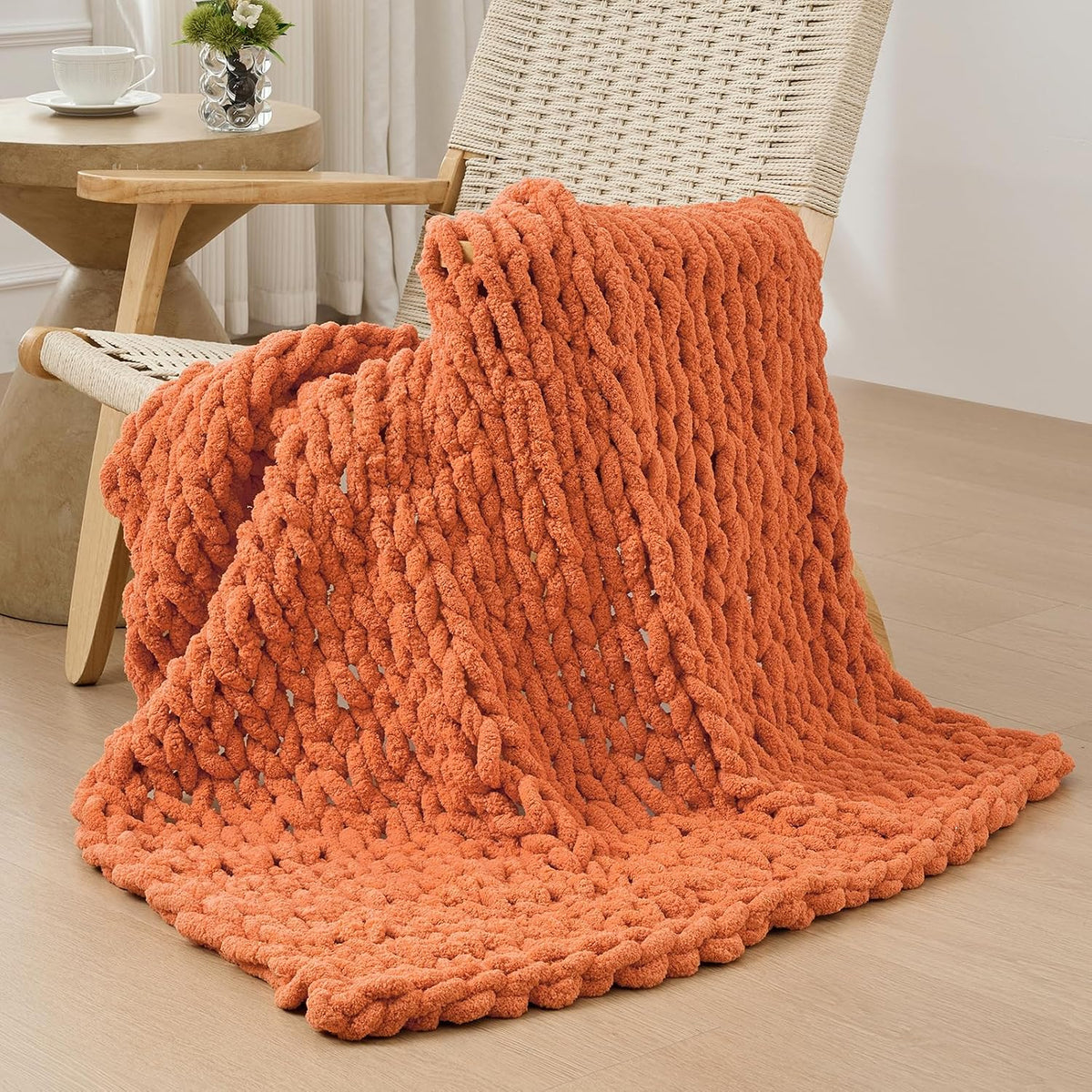 Rust-orange chunky chenille throw draped over a woven lounge chair, pooling onto a pale wood floor.
