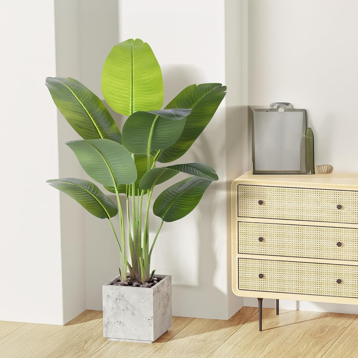 Bird of Paradise faux plant in a square concrete planter beside a rattan-front dresser in a living room setting