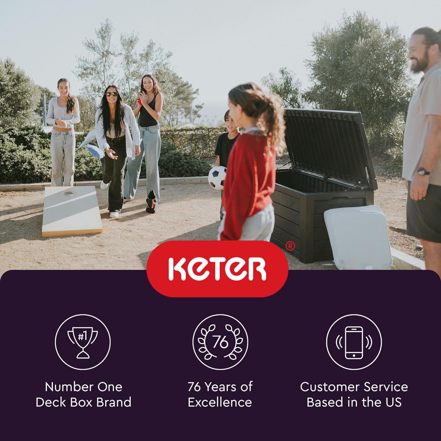 Outdoor scene with people playing cornhole next to an open dark plastic storage chest with cushions nearby.