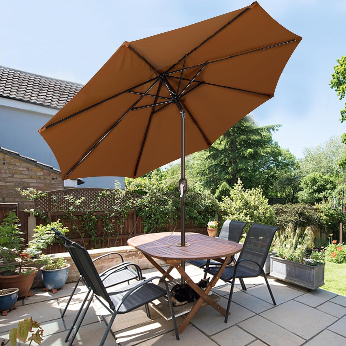 Brown outdoor umbrella over a wooden patio table and four chairs in a garden setting.