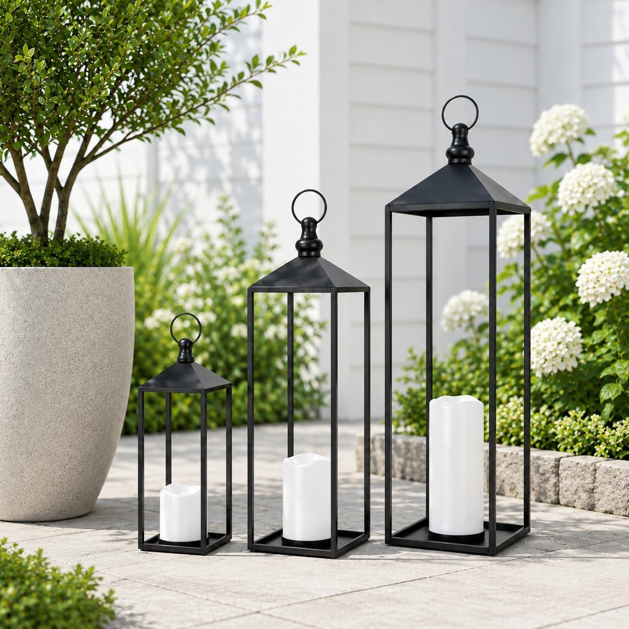 Three black metal lanterns on a sunlit patio with pavers, a tall planter, and white flowering shrubs.