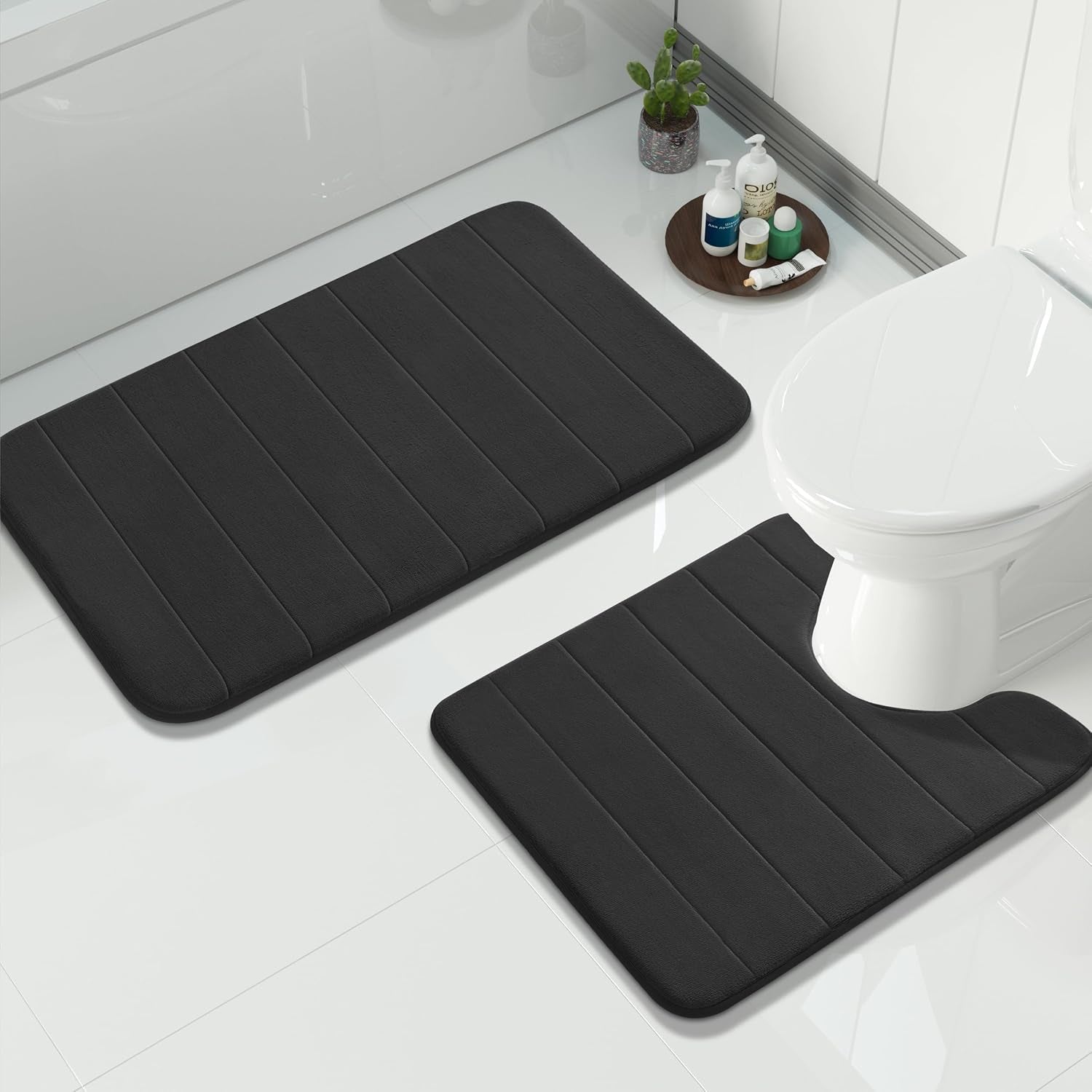 Black memory-foam bath mats on white tile, one U-shaped placed around a toilet, with toiletries on a tray nearby.