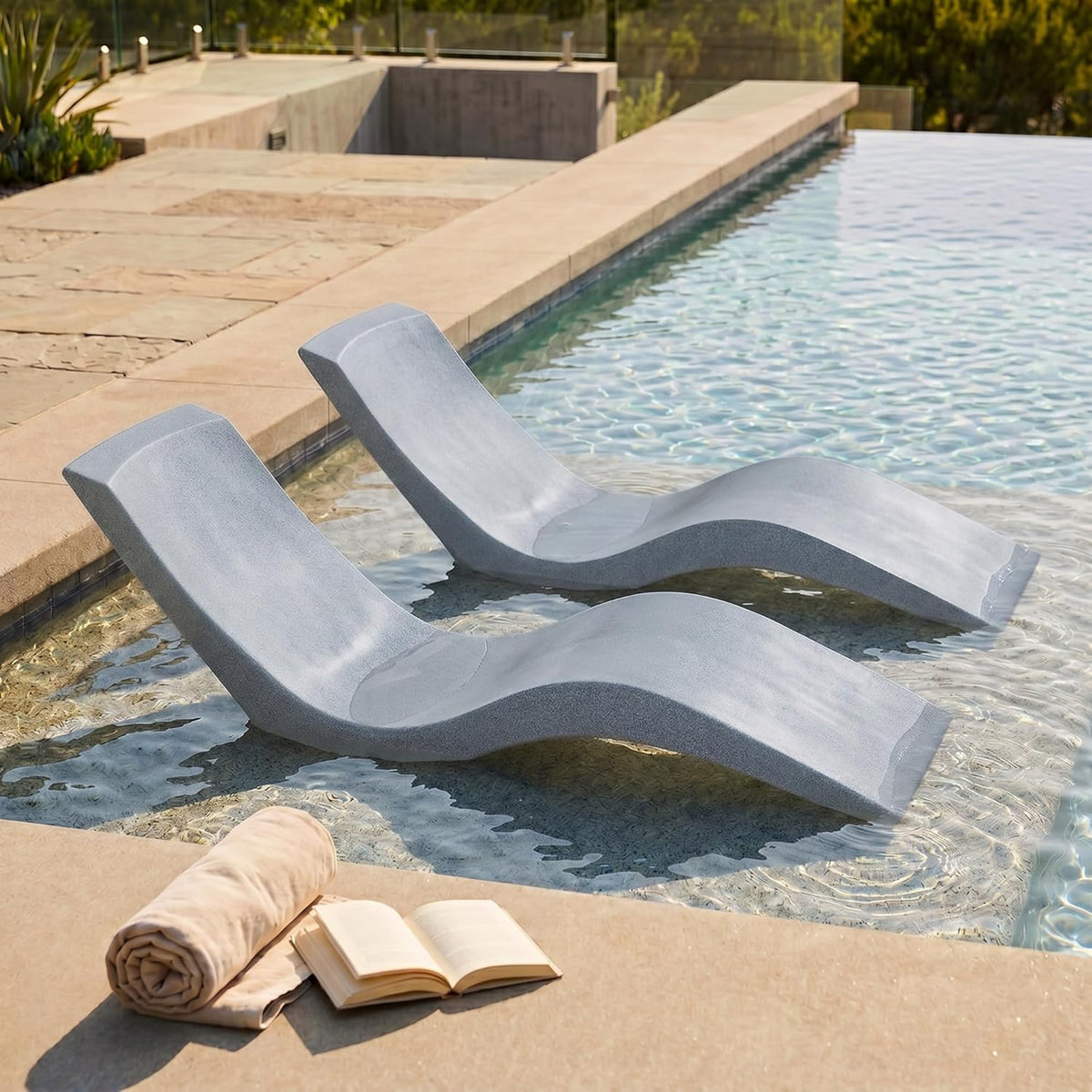 Two gray contoured loungers resting in shallow pool water near a tiled coping with a towel and book on the deck.
