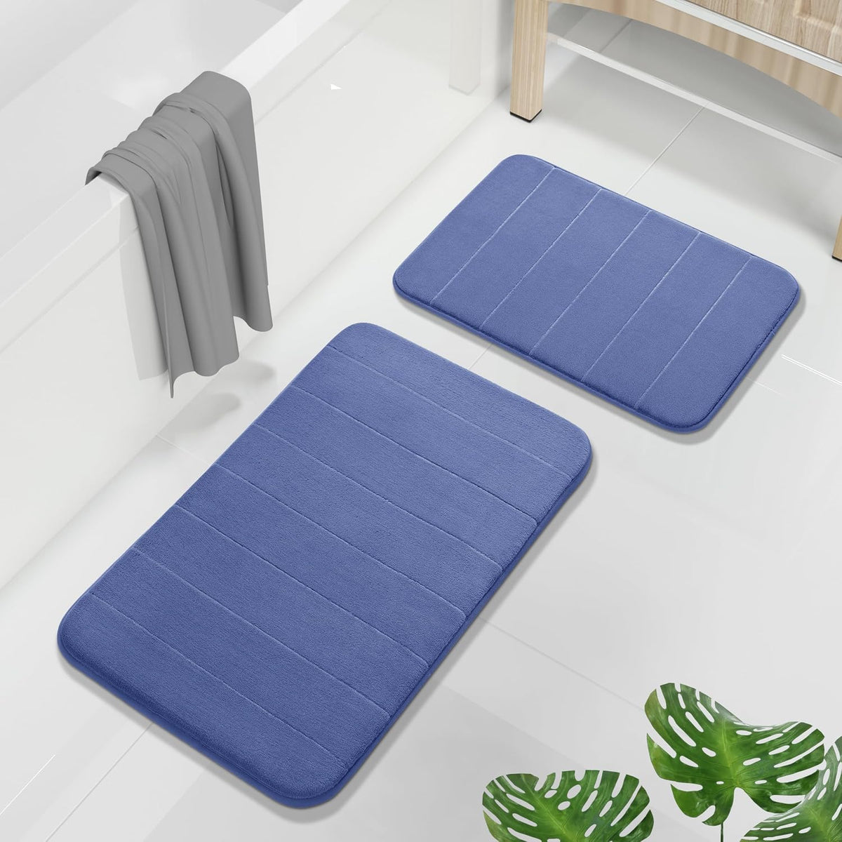 Two blue memory foam bath mats arranged on a white tiled floor next to a bathtub and wooden stool.