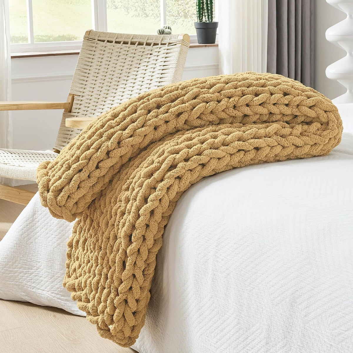 Beige chunky knit throw folded at the edge of a white bed with a woven chair in the background