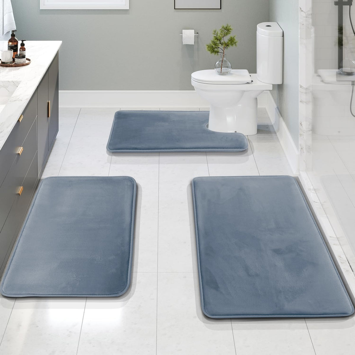 Stone blue bath rugs on white tile floor with toilet and vanity, angled bathroom view showing contour mat