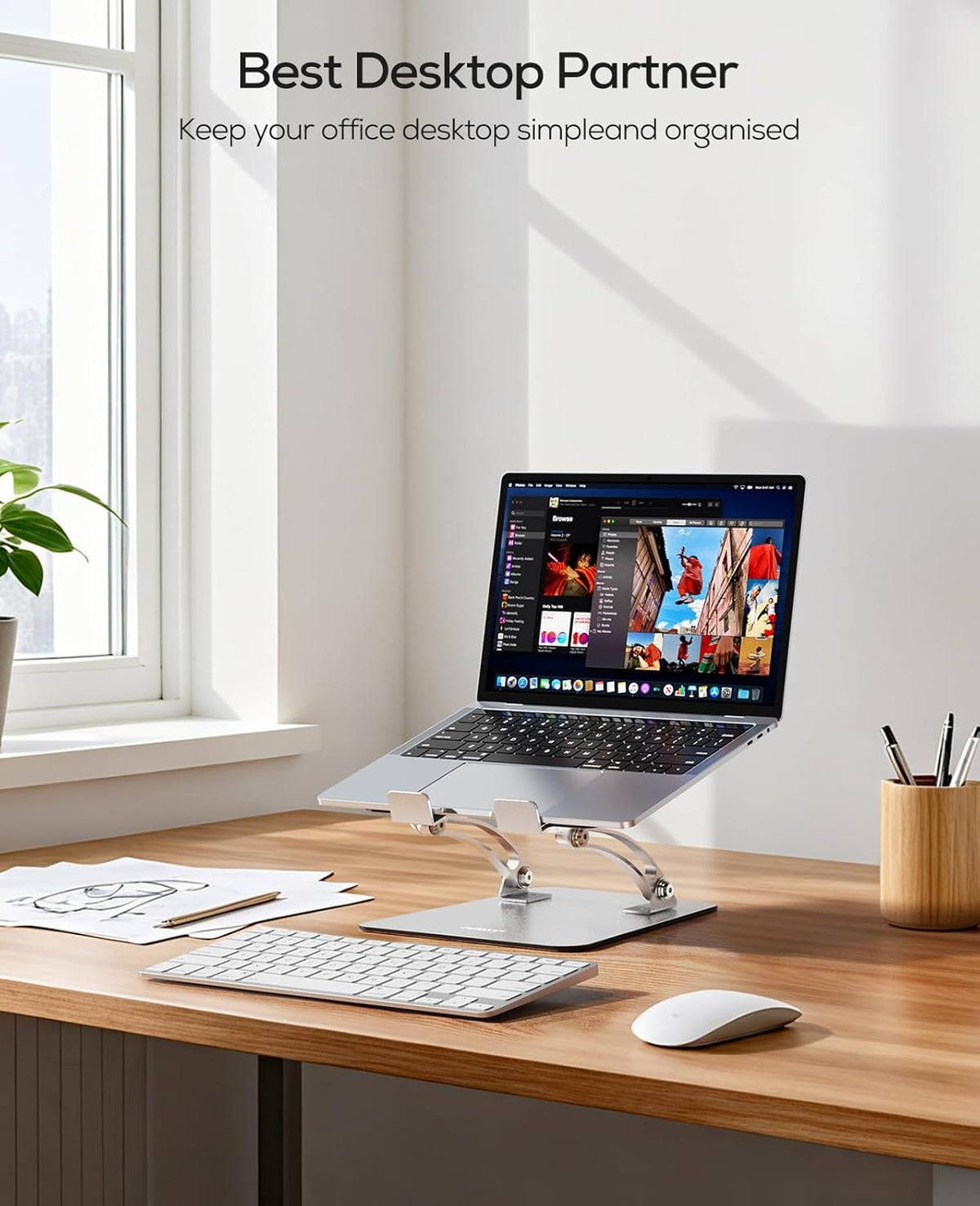 Desk scene with a raised laptop on the stand, wireless keyboard and mouse in the foreground and sunlight from a nearby
