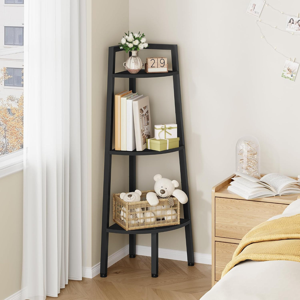 Angled corner view of a black ladder shelf with a woven basket of teddy bears on the lower shelf and a bedside table with an