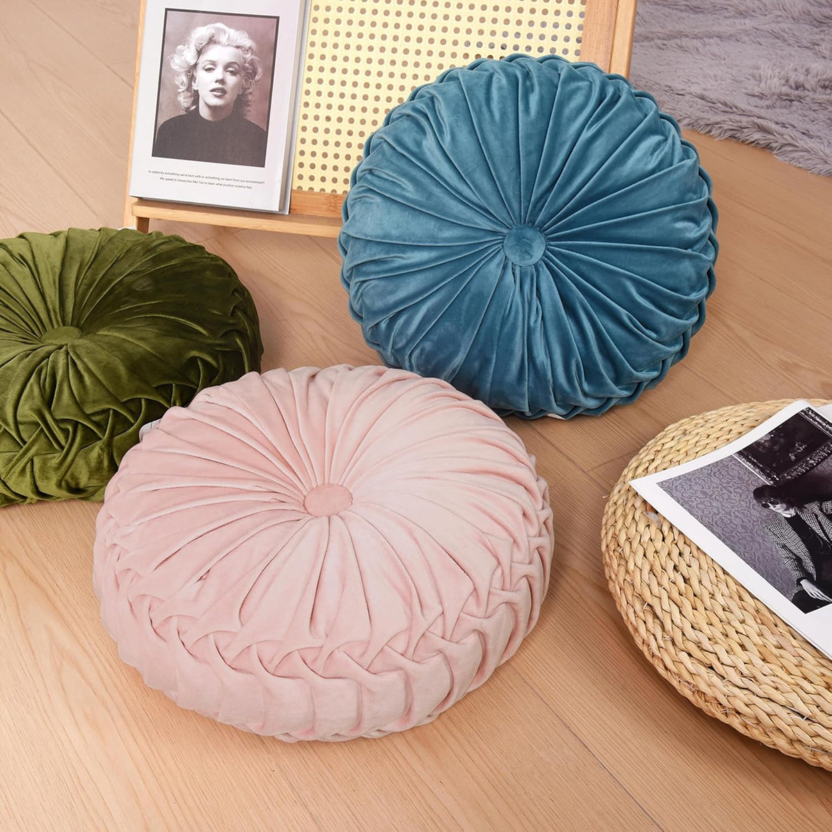 Dusty blue and olive green velvet ruched round pillows with a pale pink pillow on a wood floor near a woven tray and magazine