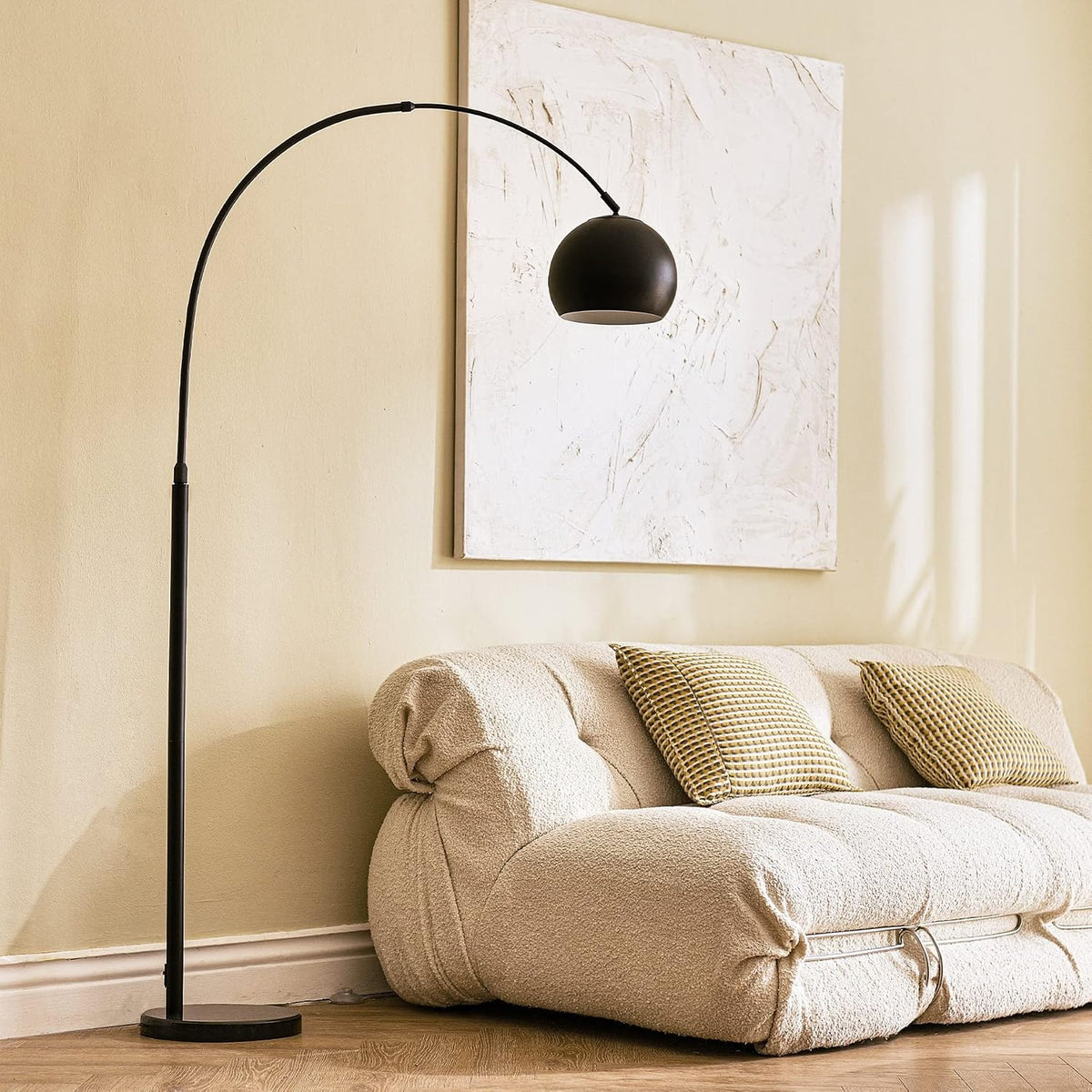 Matte black arc lamp arching over a cream sofa with textured pillows and abstract wall art