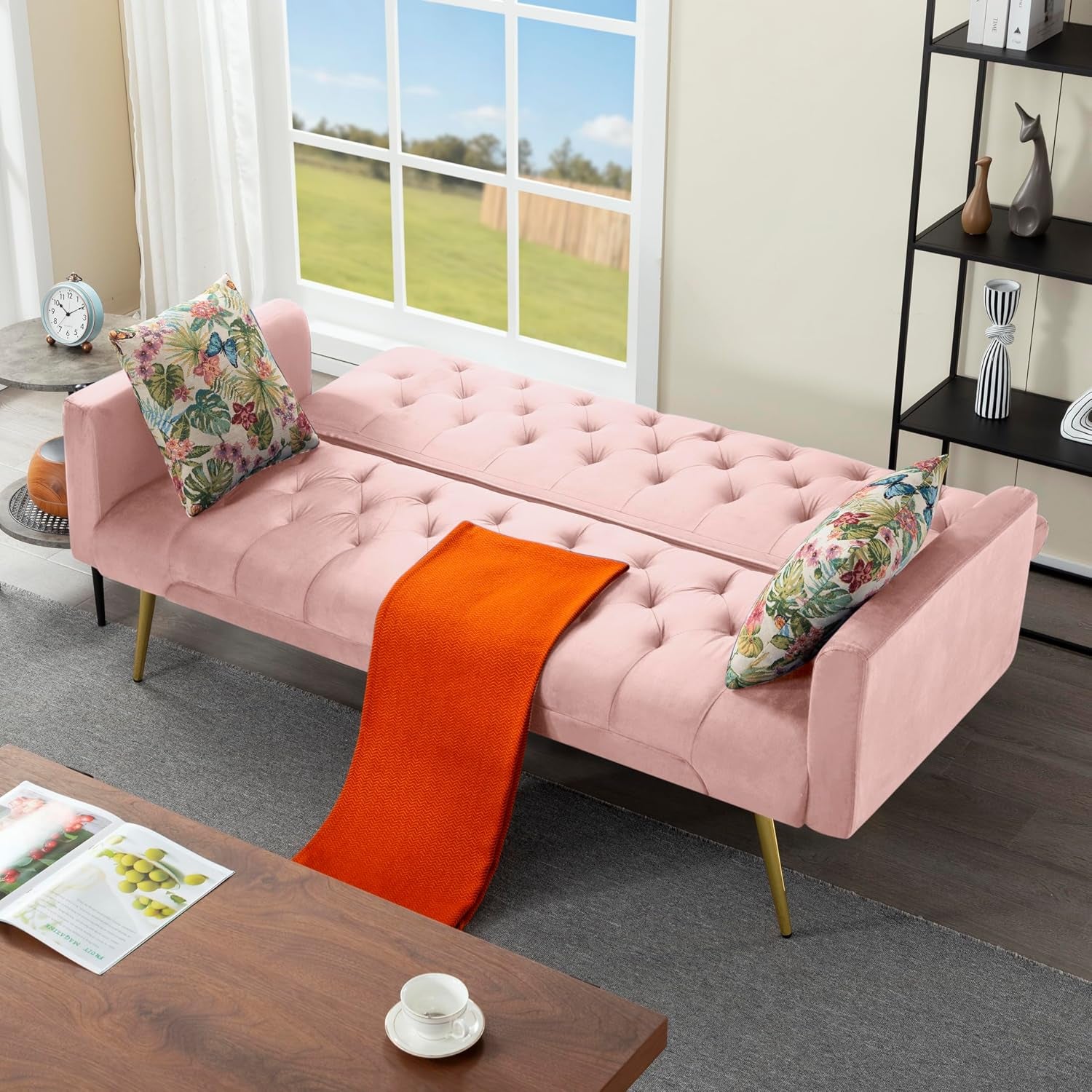 Pink velvet futon shown flat as a bed with two floral throw pillows and an orange throw.
