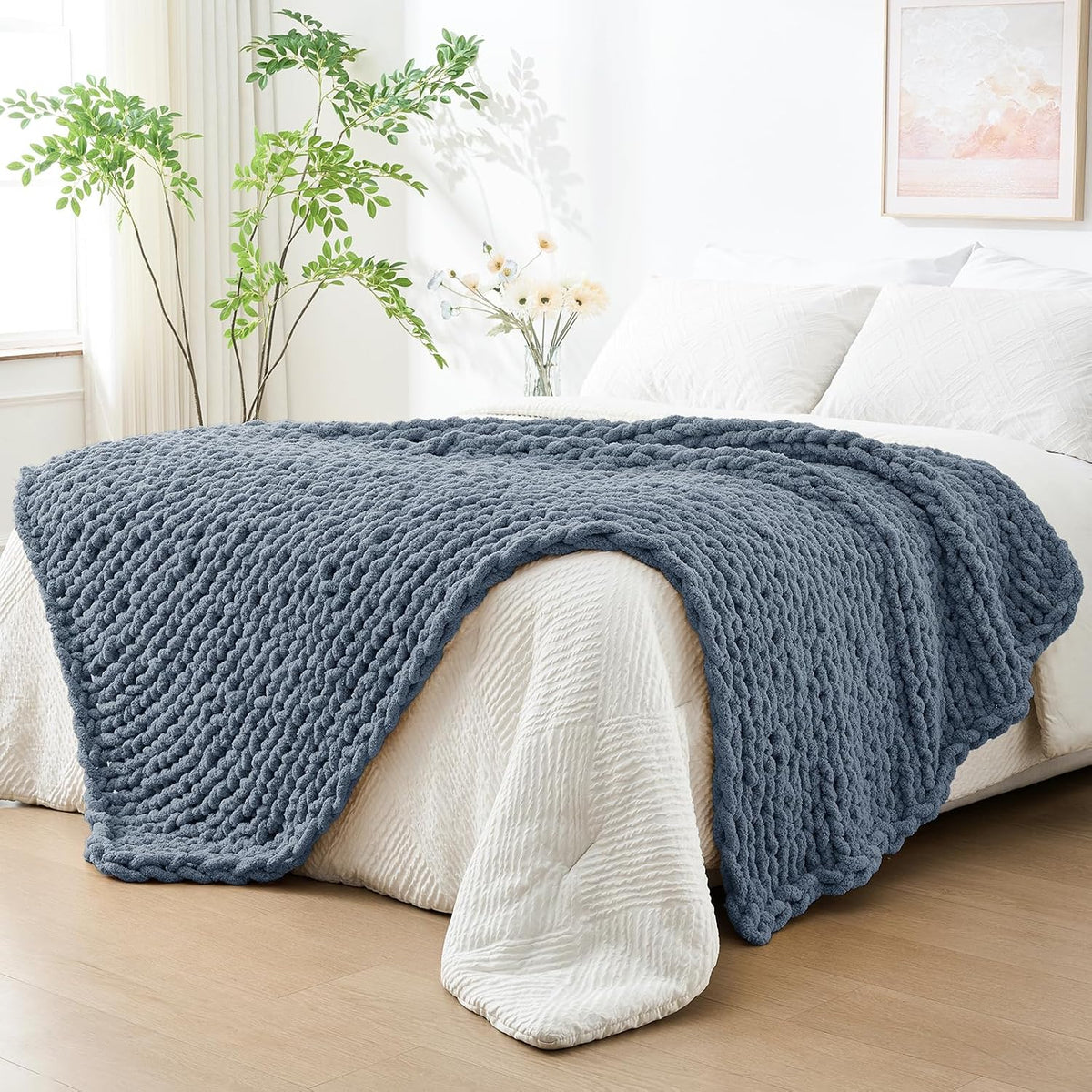 Slate blue chunky knit throw laid over the foot of a bed with plants and framed art in background