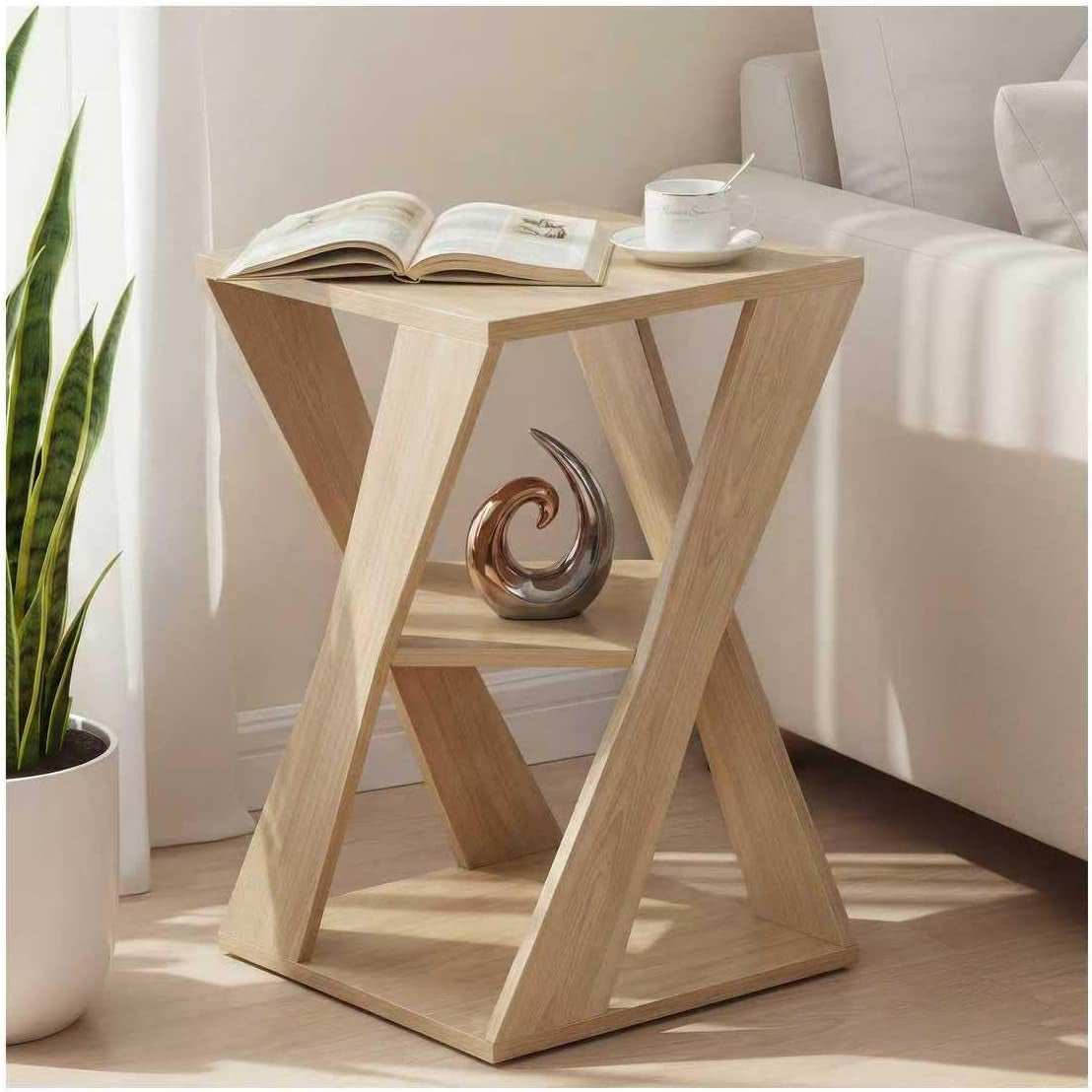 Light wood X-shaped end table beside a sofa with a book and cup on top and decorative sculpture on the middle shelf.
