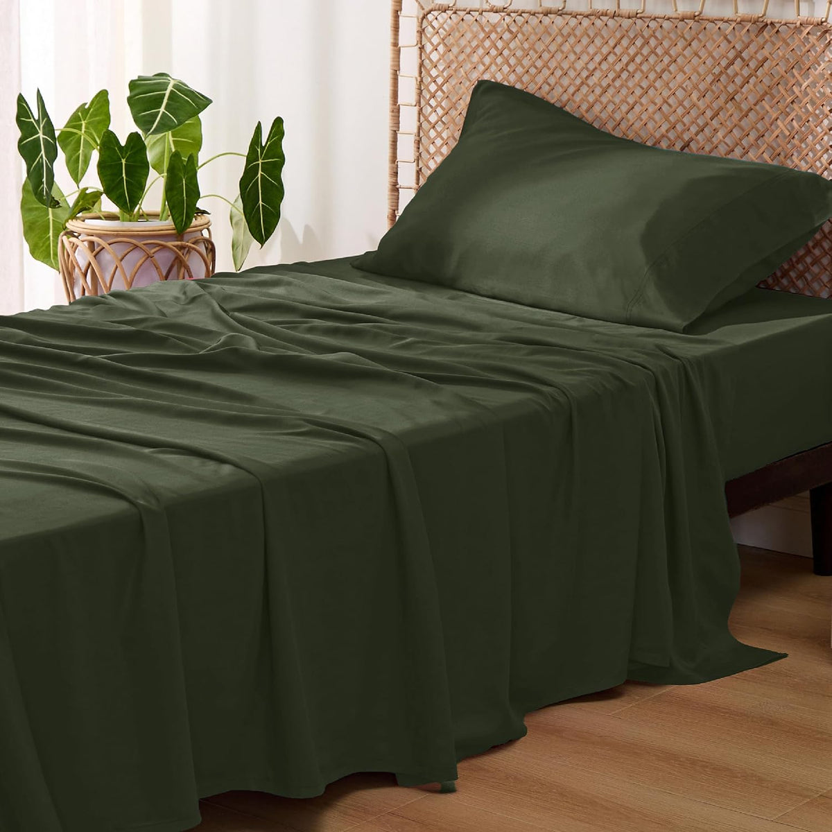 Angled view of a bed with olive green sheets and a pillow, a potted plant beside a woven headboard.