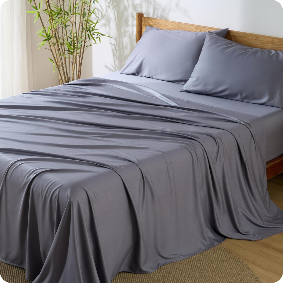 Charcoal gray sheets and two pillows draped over a wooden bed frame with soft natural light.