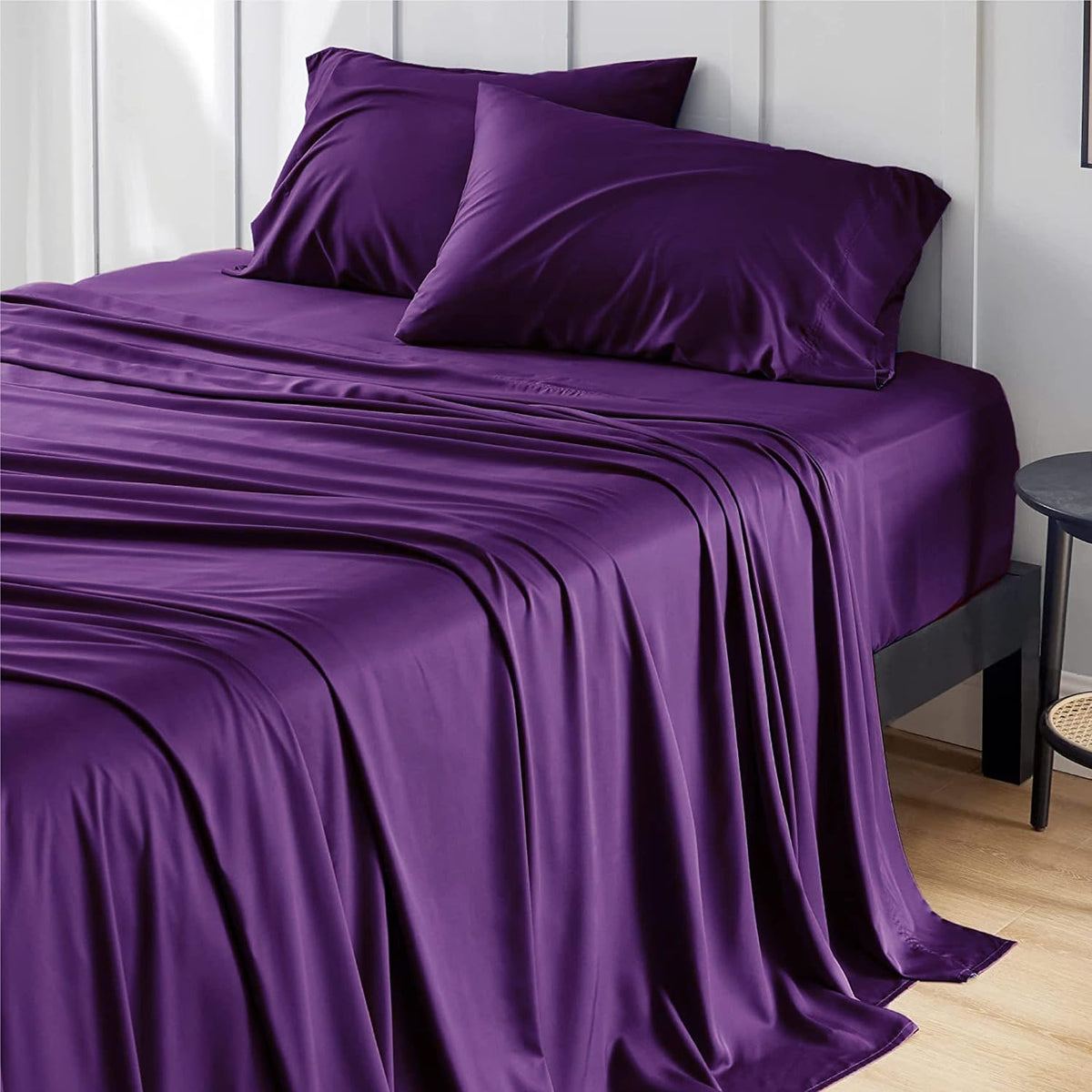 Three-quarter view of a bed with deep purple sheets and two matching pillows, sheets draping to the floor.