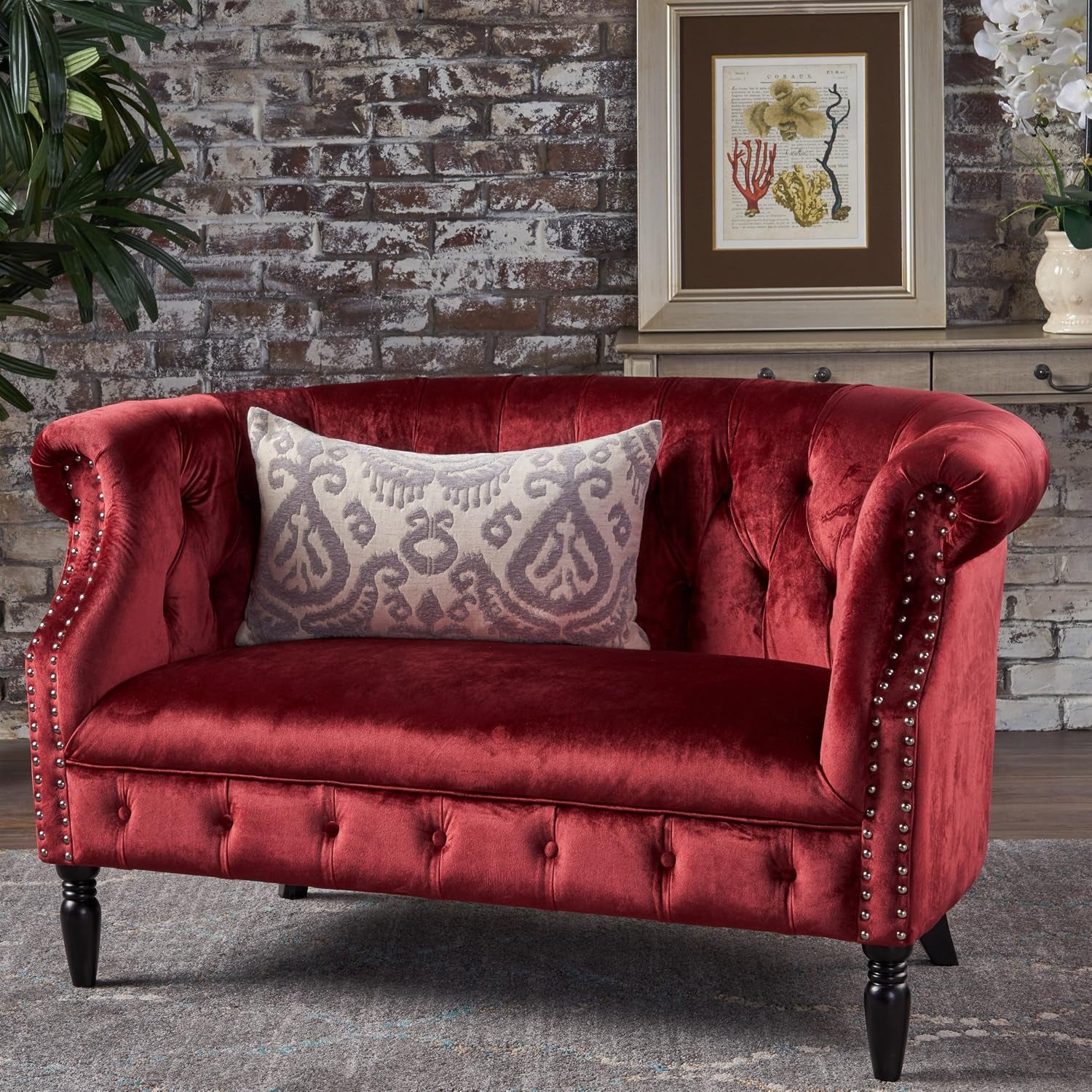 Styled room scene with red velvet tufted loveseat, patterned lumbar pillow, brick wall and framed art