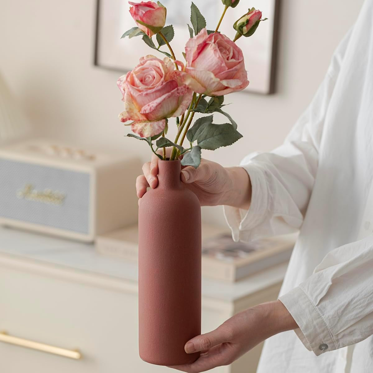 Hands holding a tall terracotta matte ceramic vase with pink roses, close-up of narrow neck and finish