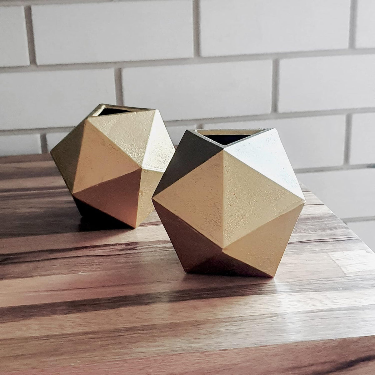 Two gold geometric cast-iron balls with open tops placed on a wood tabletop in front of white subway tile.