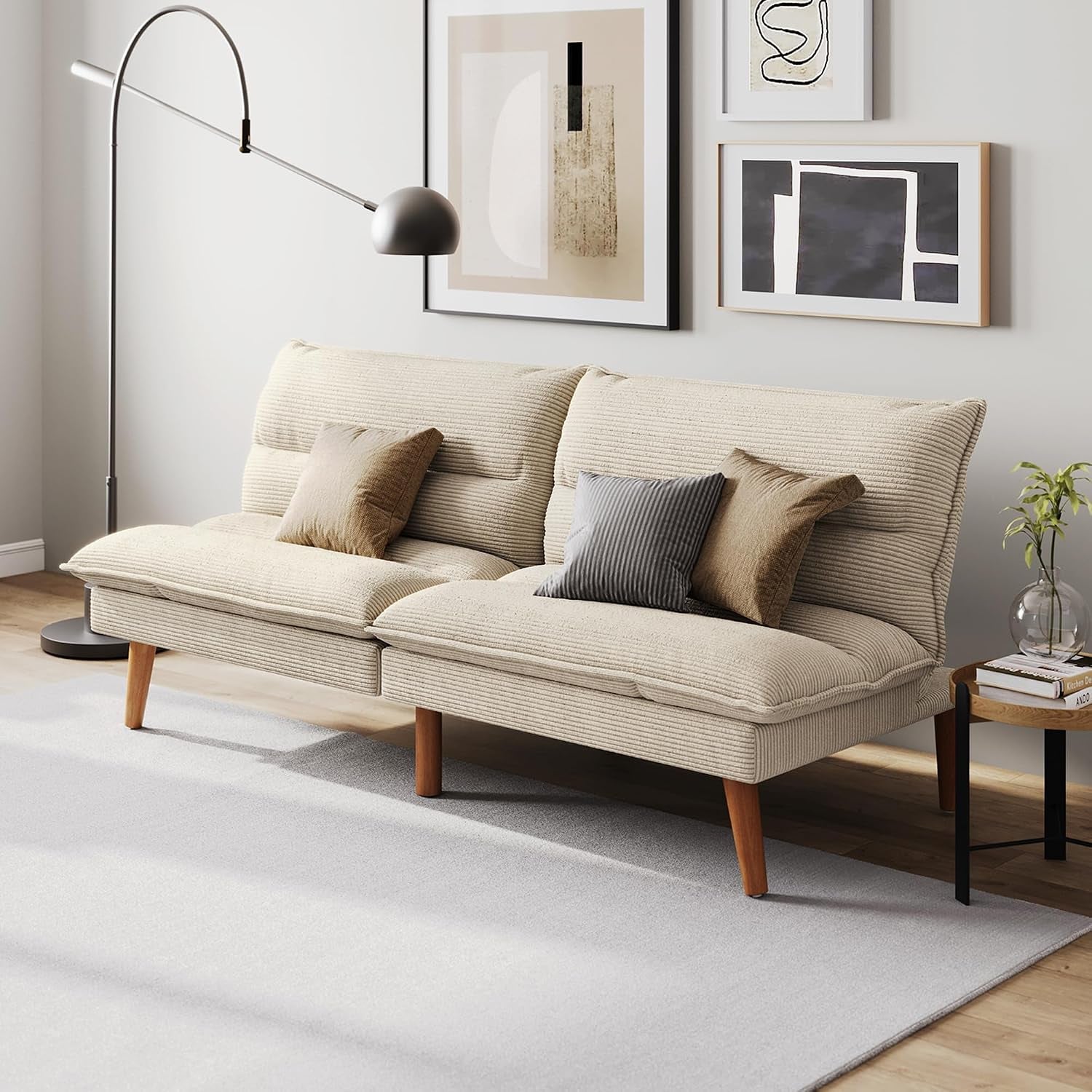 Beige corduroy convertible loveseat with wooden legs, three throw pillows, floor lamp, art, and side table.