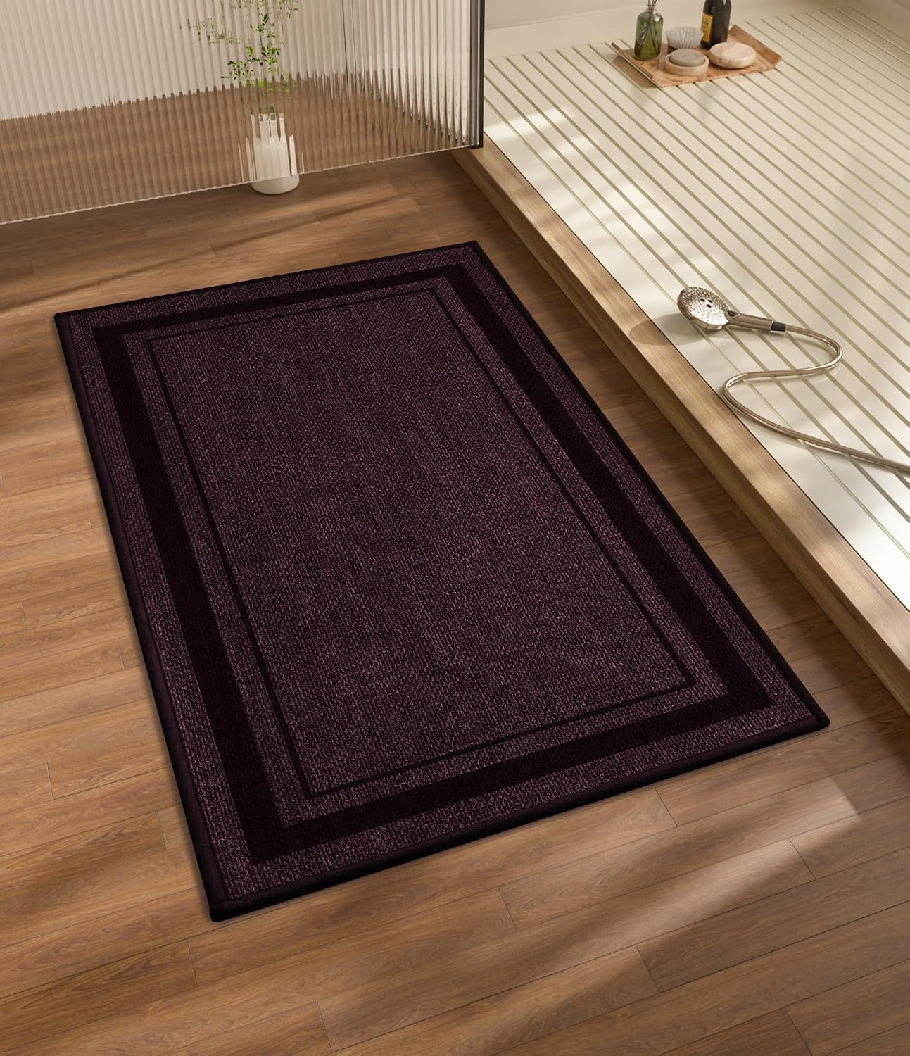 Rectangular dark purple bath rug with double border placed on wood floor beside a tub area.