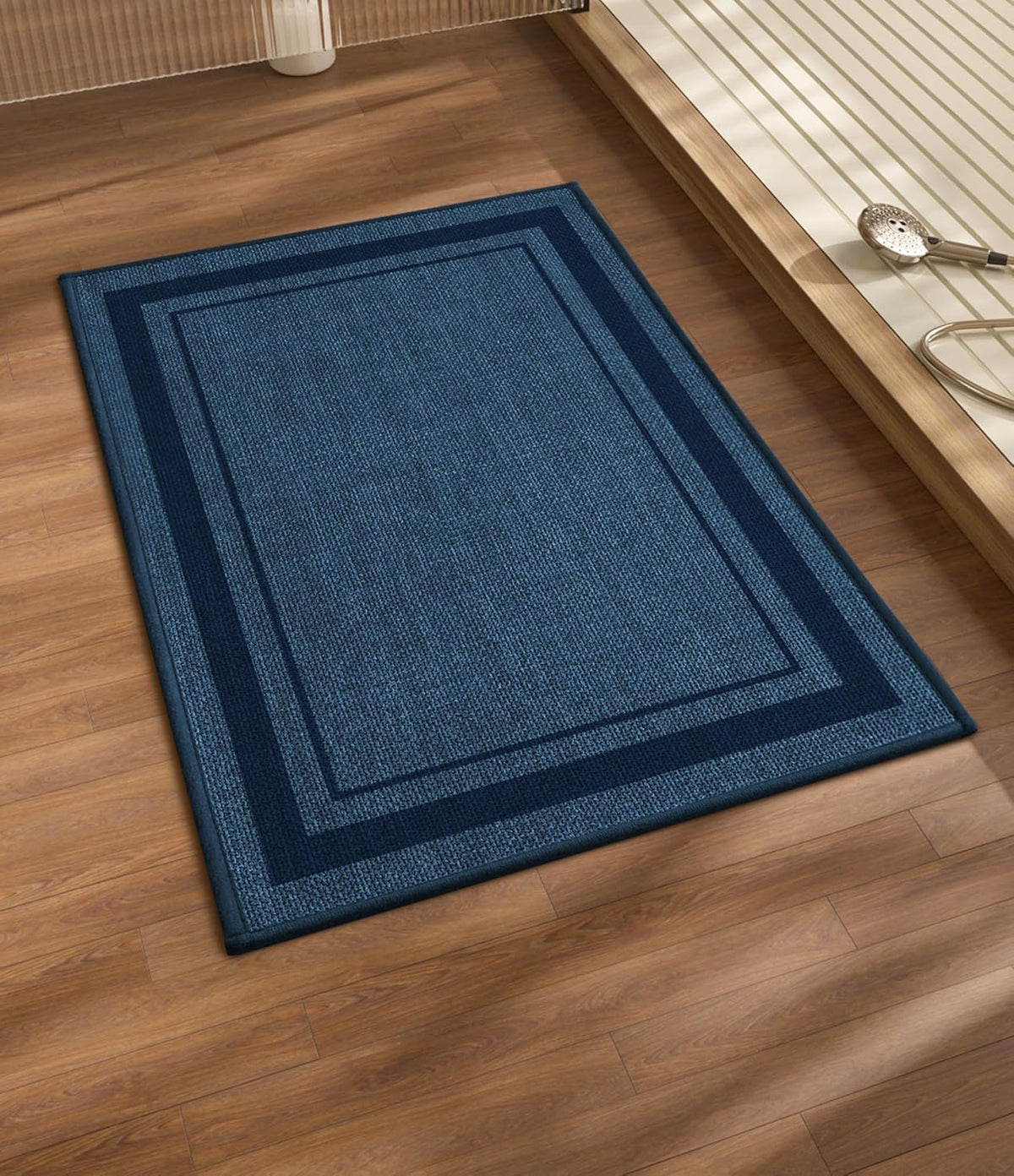 Blue rectangular bath mat with contrasting darker double-border on wood flooring near a tub.
