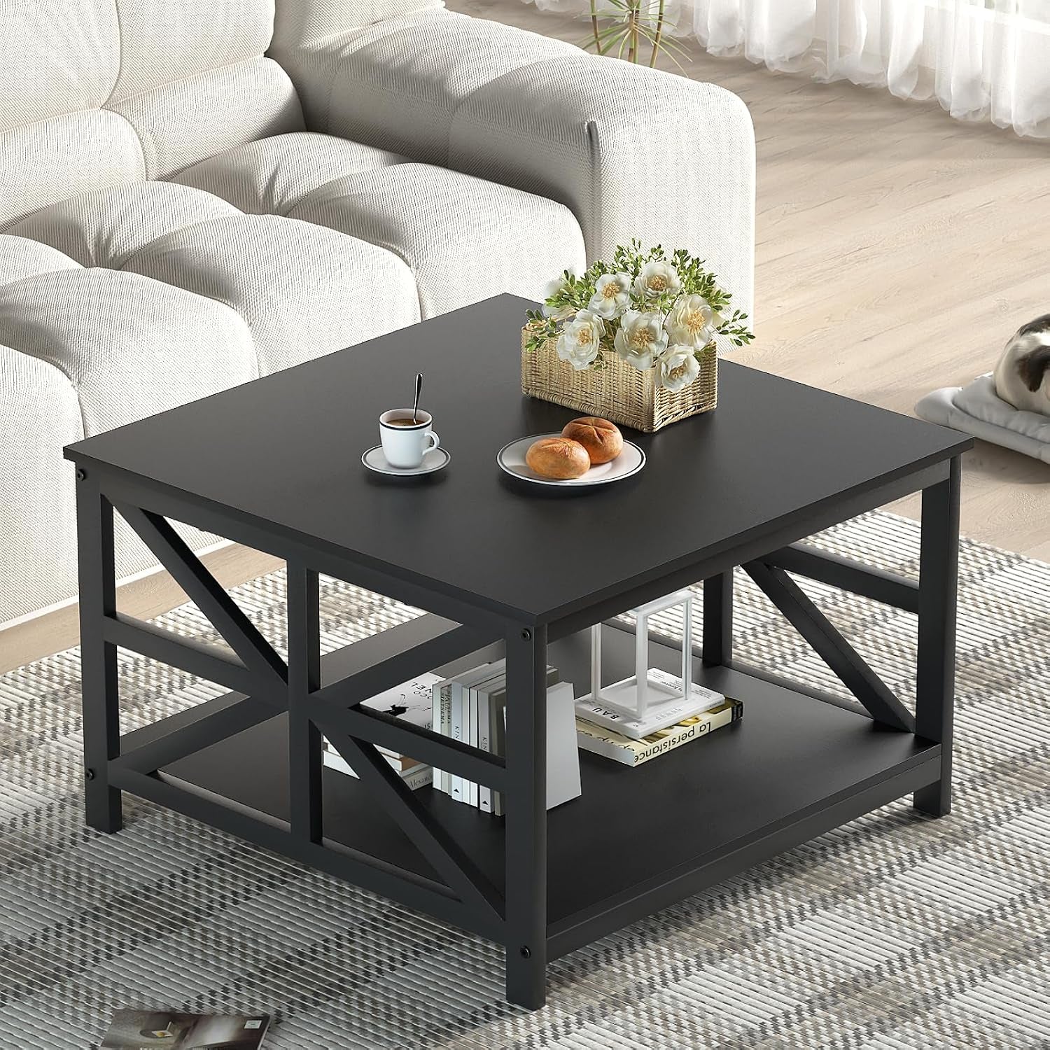Square black coffee table with X-frame sides and lower shelf beside a light sofa, styled with flowers and pastries.