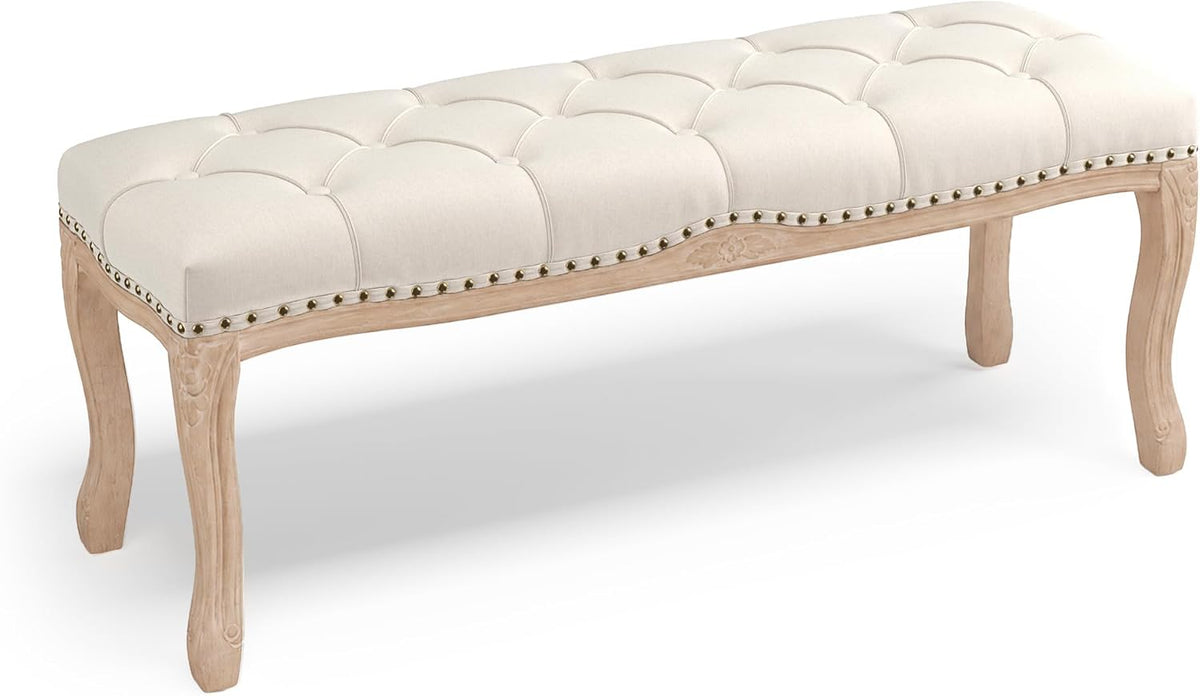 Beige linen diamond-tufted bench angled front-left view showing carved wooden legs and brass nailhead trim