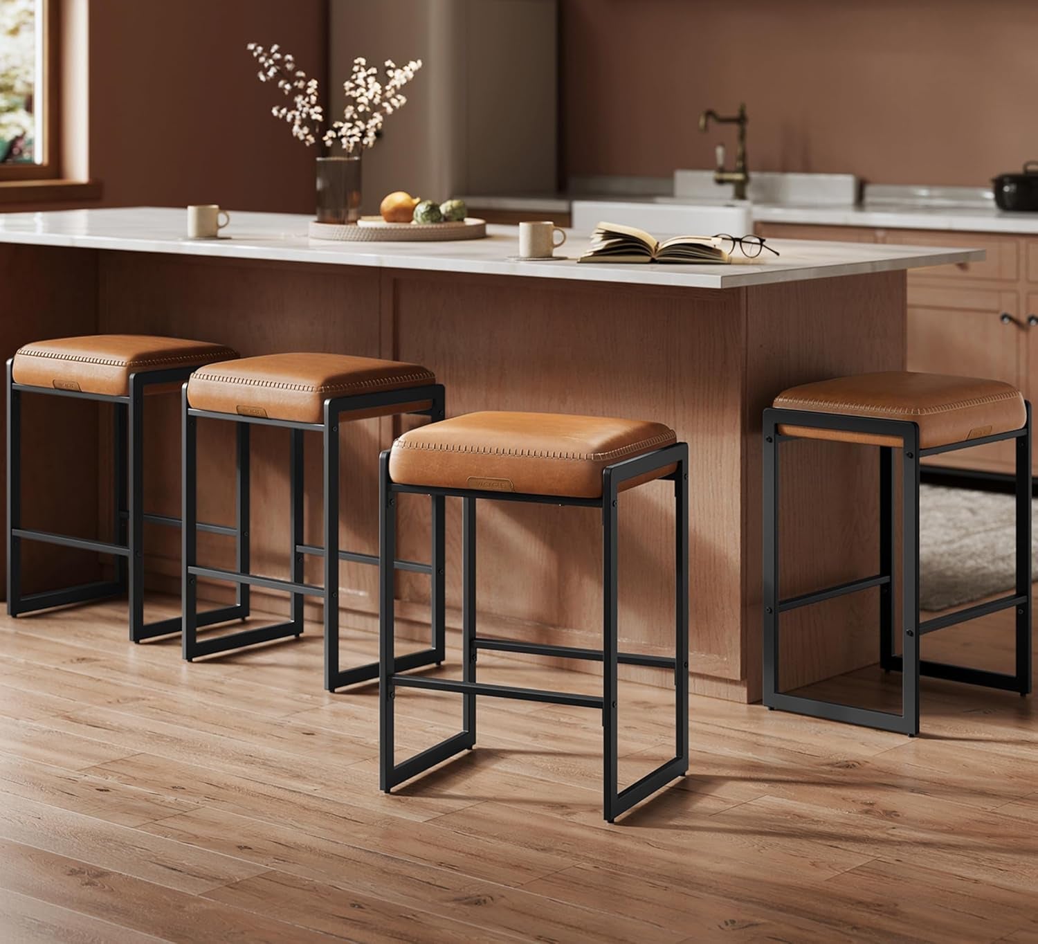 Four caramel-cushioned stools lined along a kitchen island, one slightly pulled out, with mugs, a fruit tray and sink visible