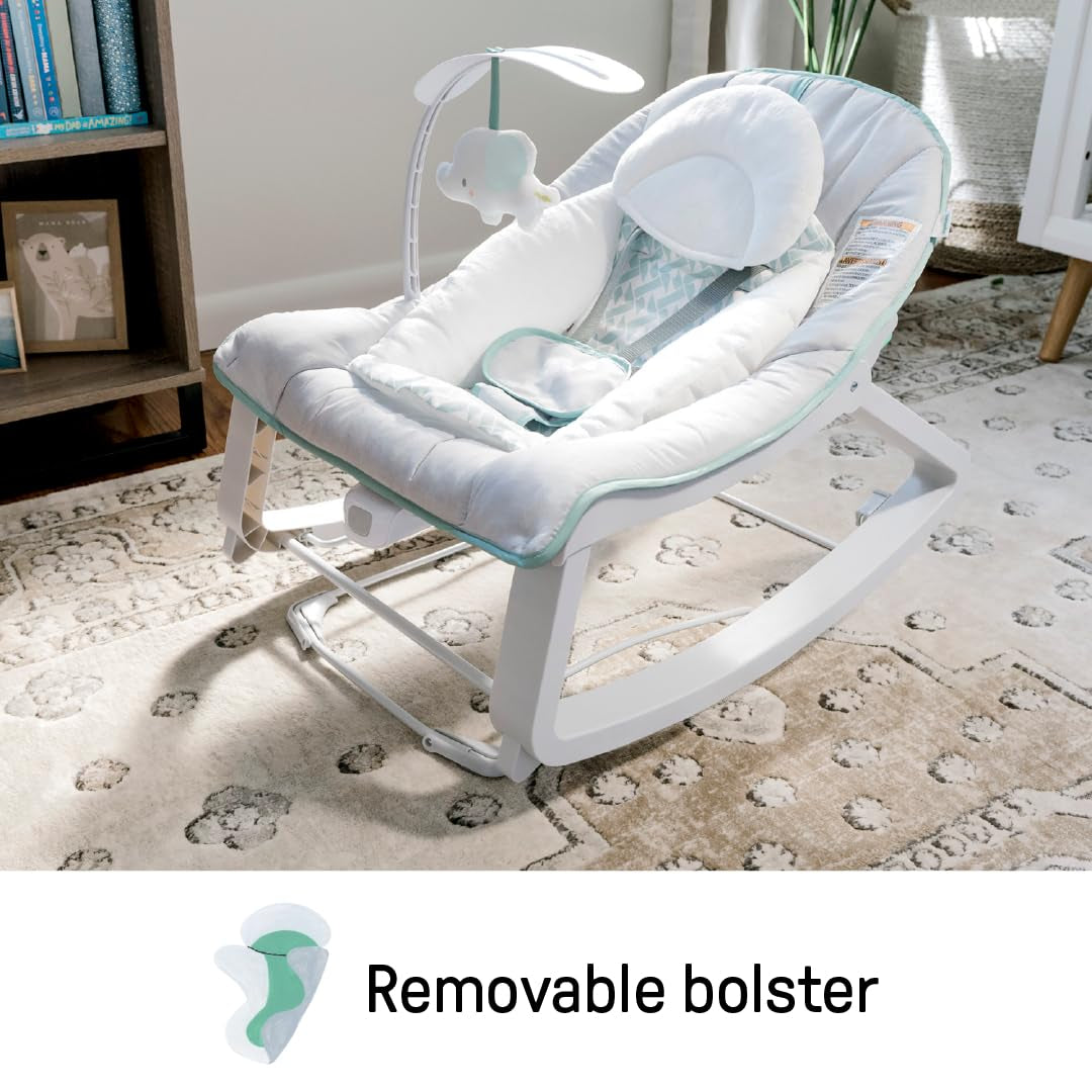 Gray and mint-trim baby bouncer placed on a patterned rug beside a bookshelf in a sunlit room.
