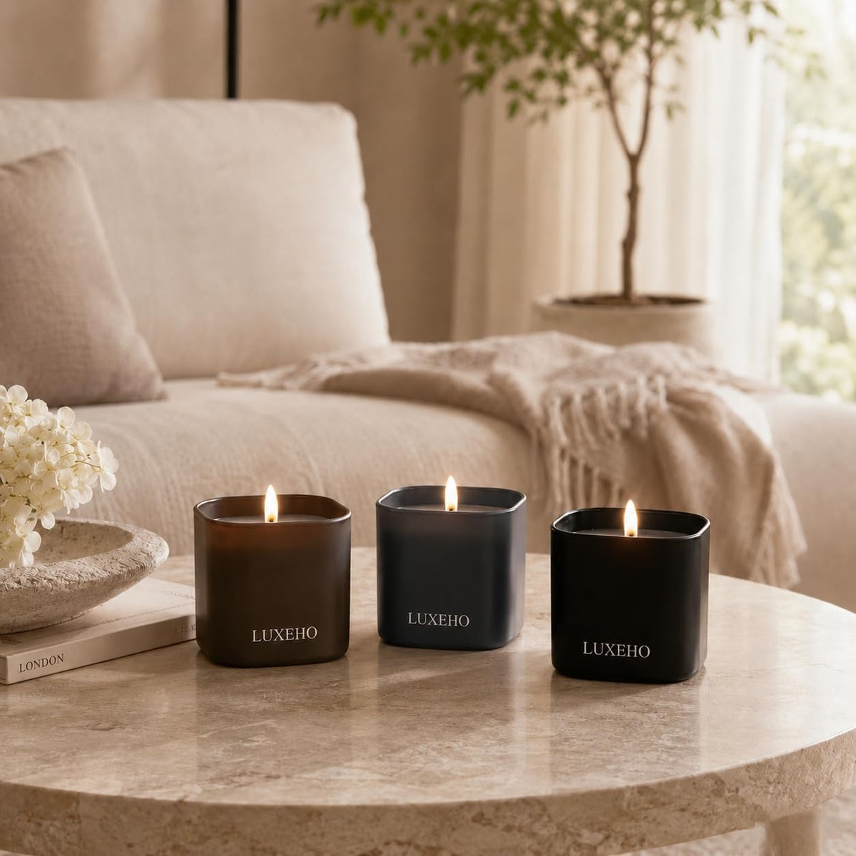 Three lit matte jars on a round stone coffee table in a softly lit living room with sofa and plant in background.