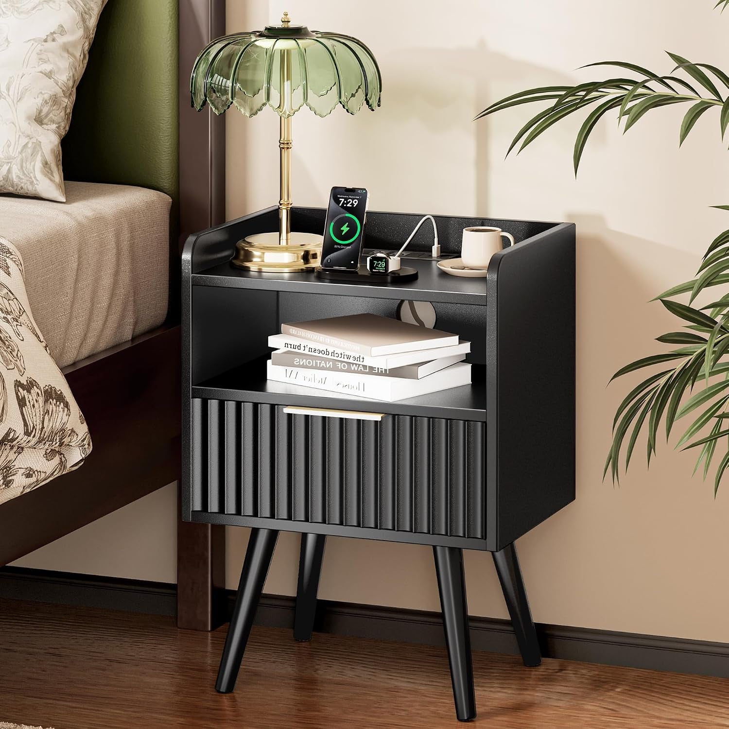 Black fluted nightstand beside bed with lamp, phone charger, mug and stacked books on open shelf.