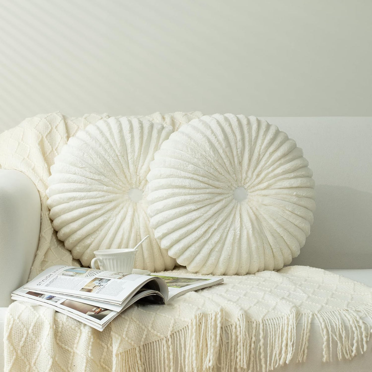 Two round ruched ivory pillows on a sofa draped with a knit throw, magazine and teacup nearby.